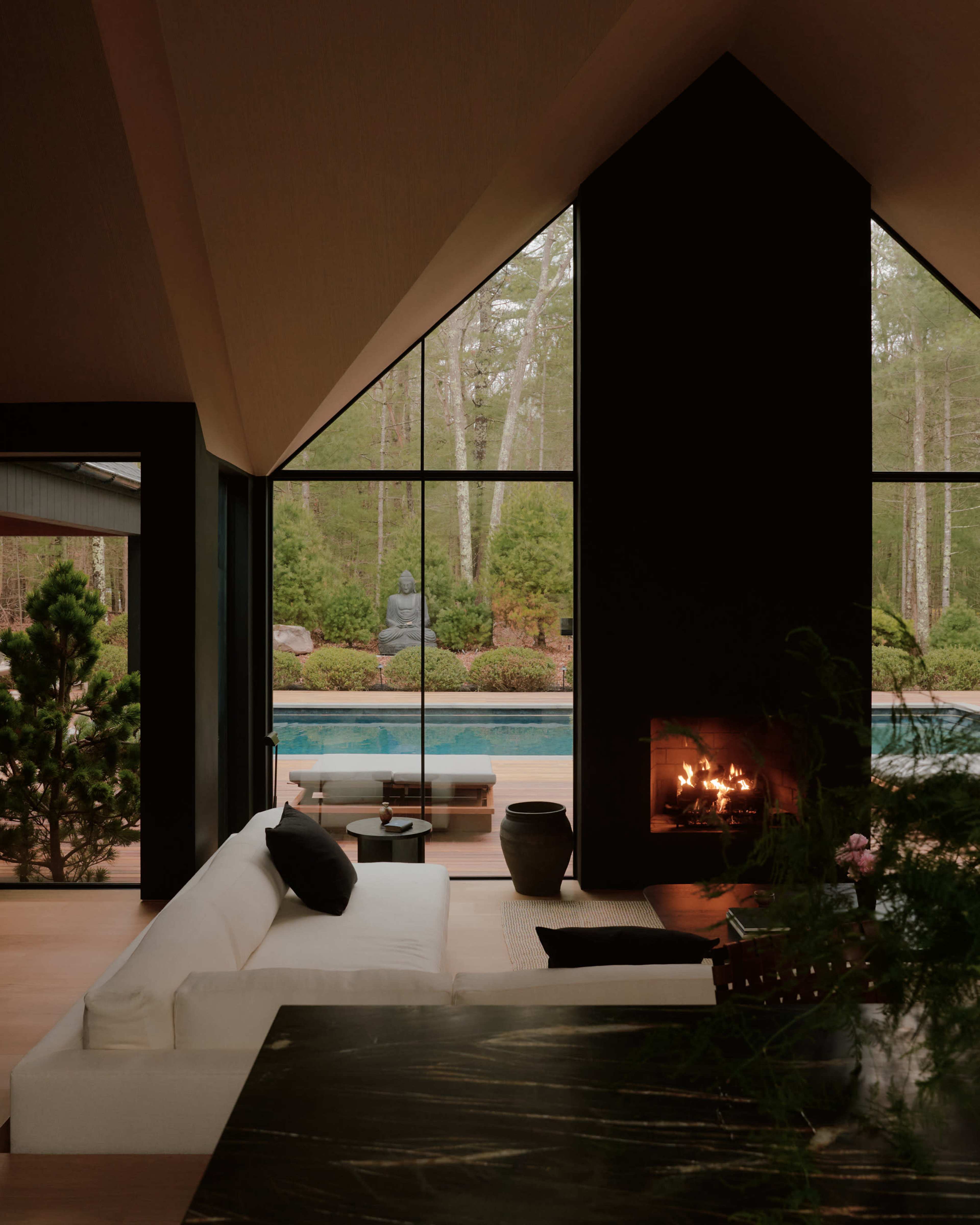 A modern living space features a geometric fireplace and large windows overlooking a garden with a statue by a pool.