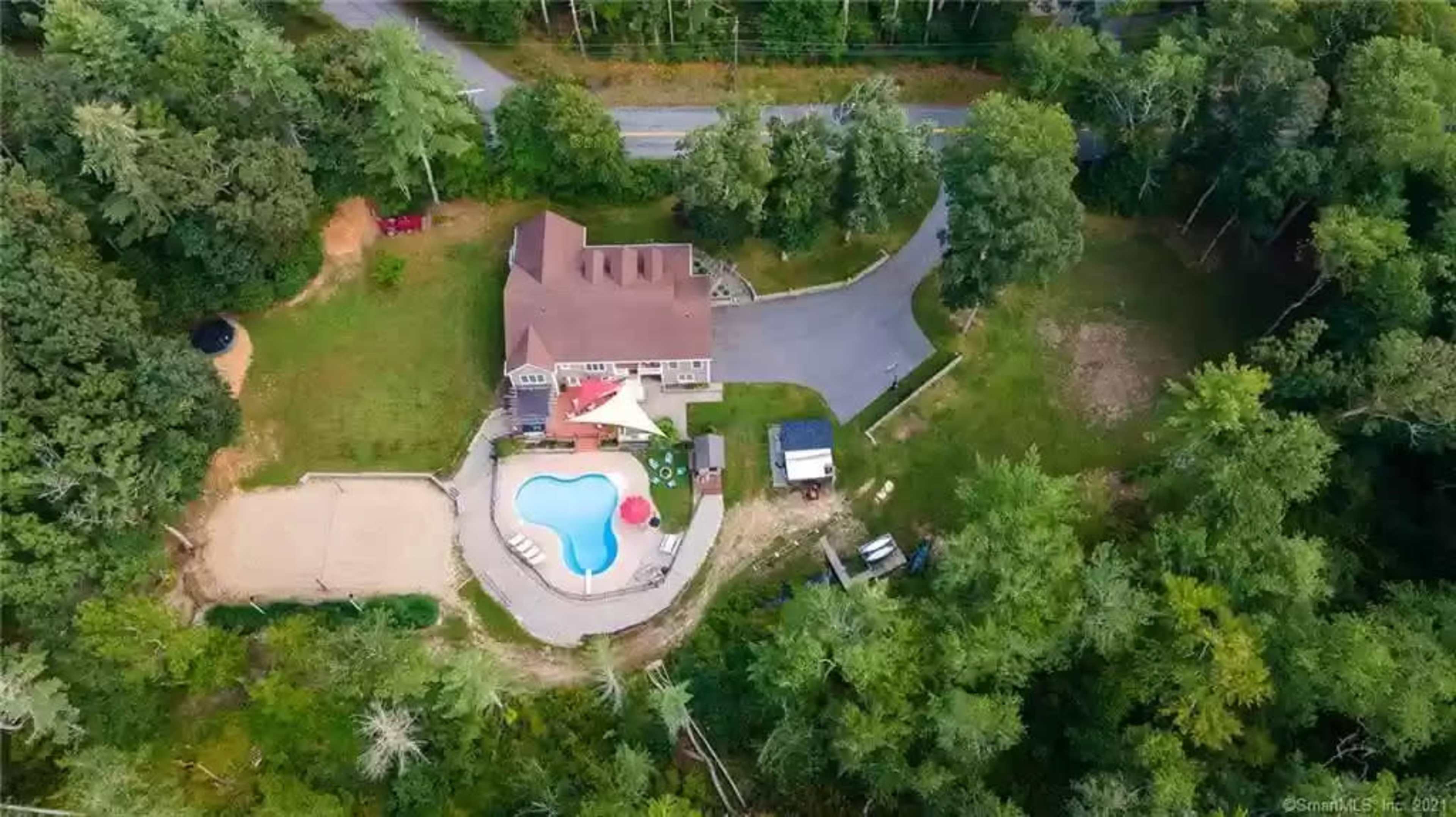 An aerial view shows a large house with a swimming pool and a sand volleyball court situated among trees near a curved road.
