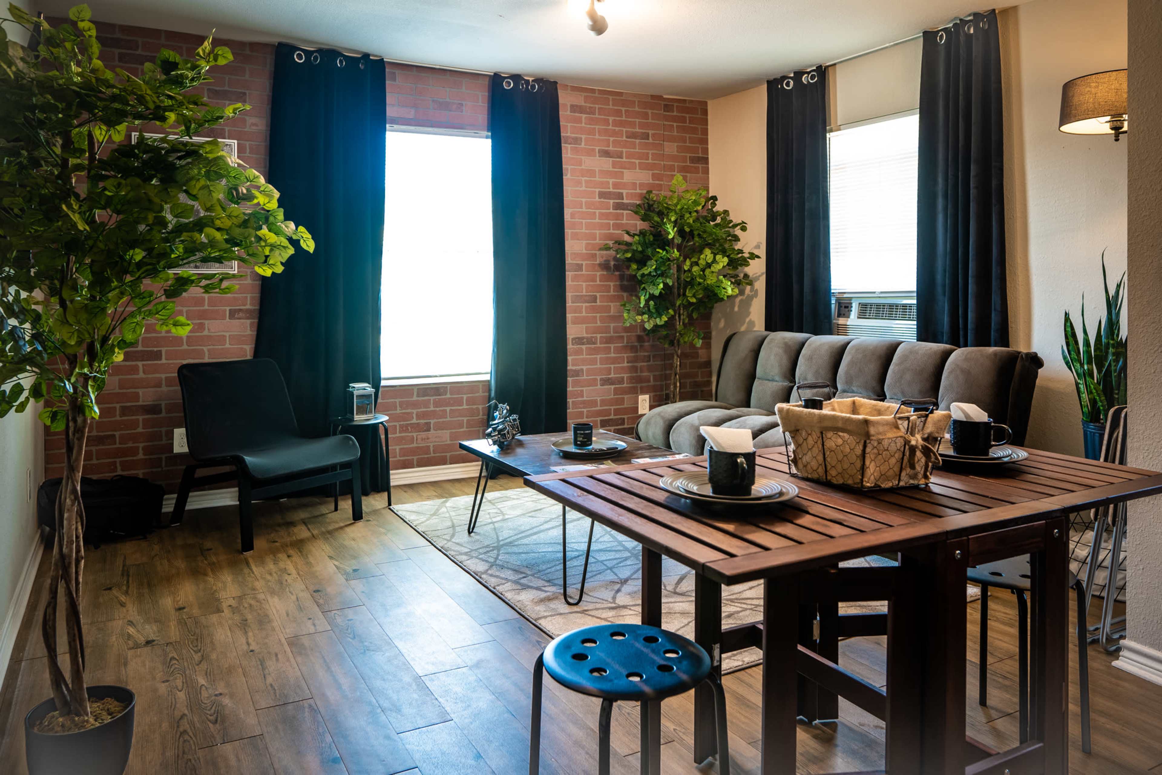 The living space features a cozy seating area with a gray couch, a wooden dining table set for four, and plants arranged against brick and windowed walls.