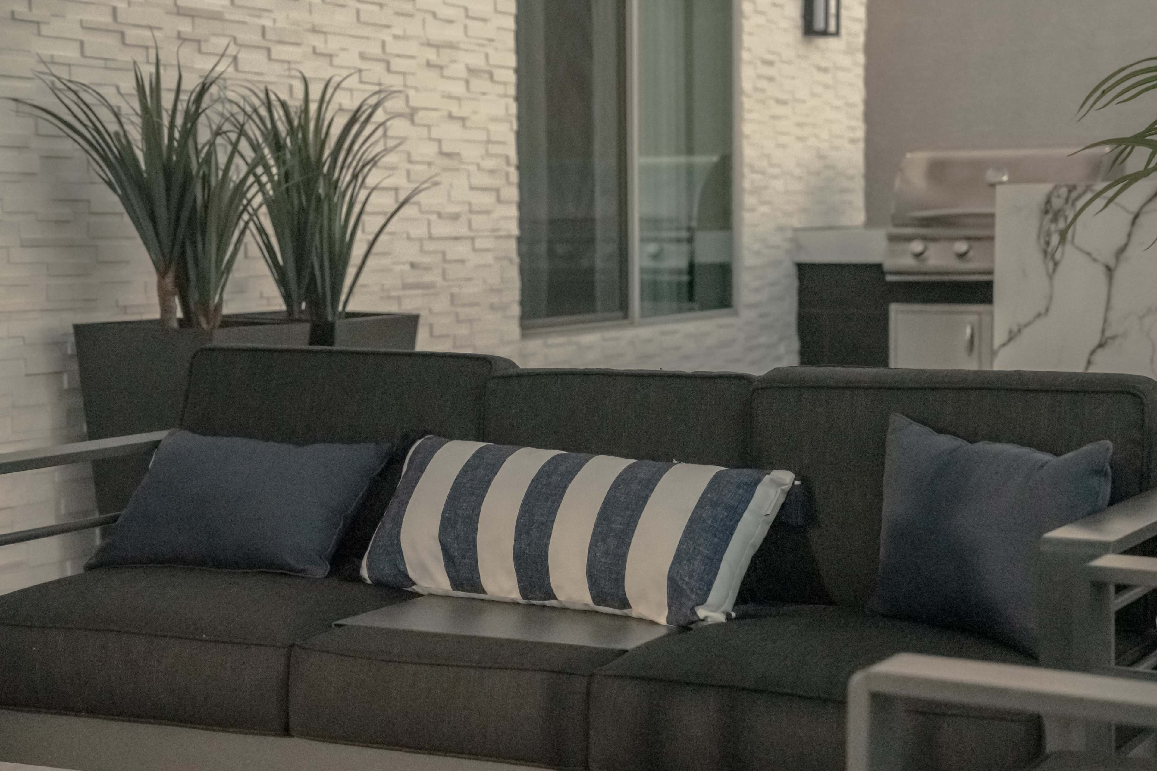 A modern outdoor seating area with a dark gray couch adorned with a striped pillow, beside potted plants and a stainless steel grill in the background.