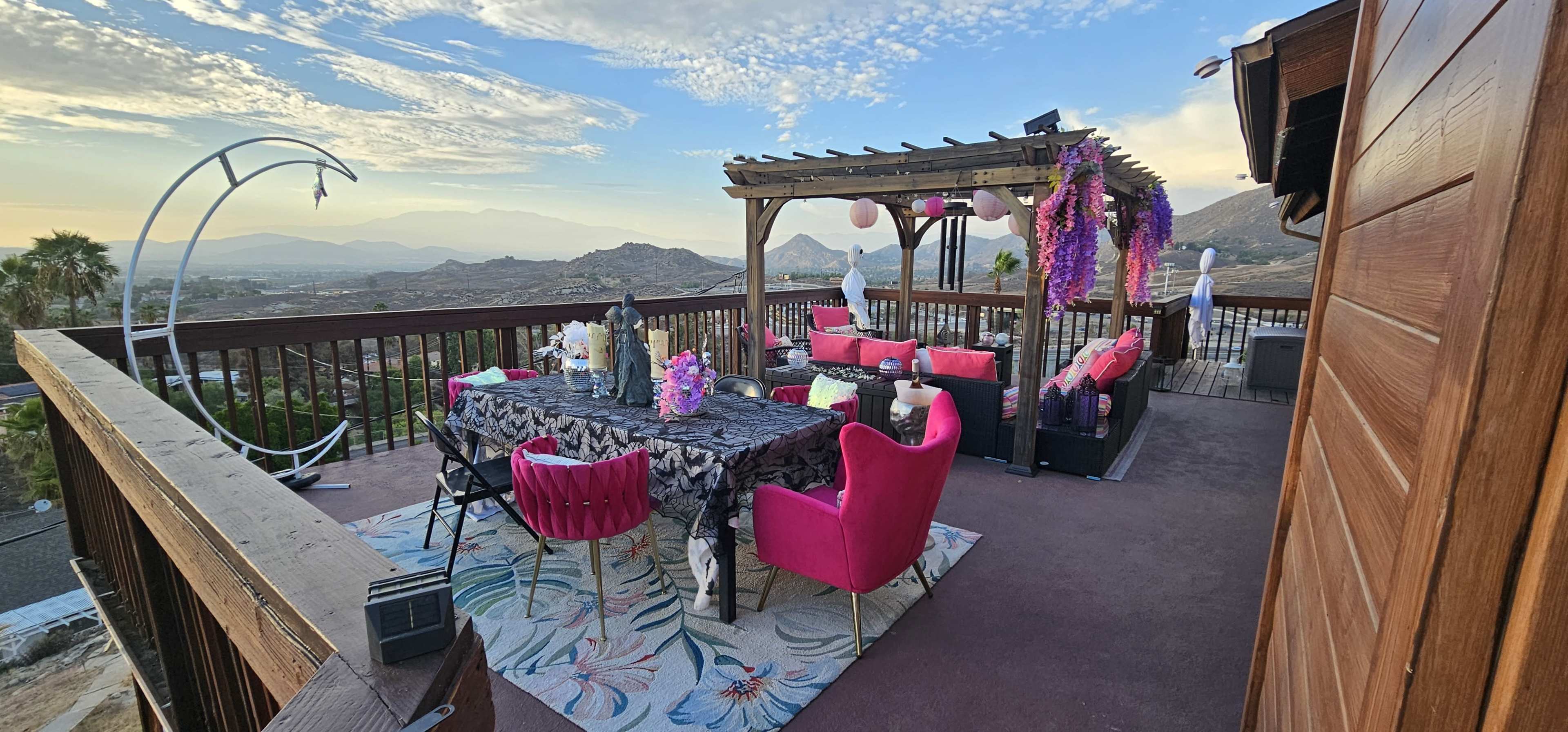 A spacious outdoor terrace features a large dining table surrounded by pink chairs, adorned with floral decorations, and offers a scenic view of hills and mountains in the background.