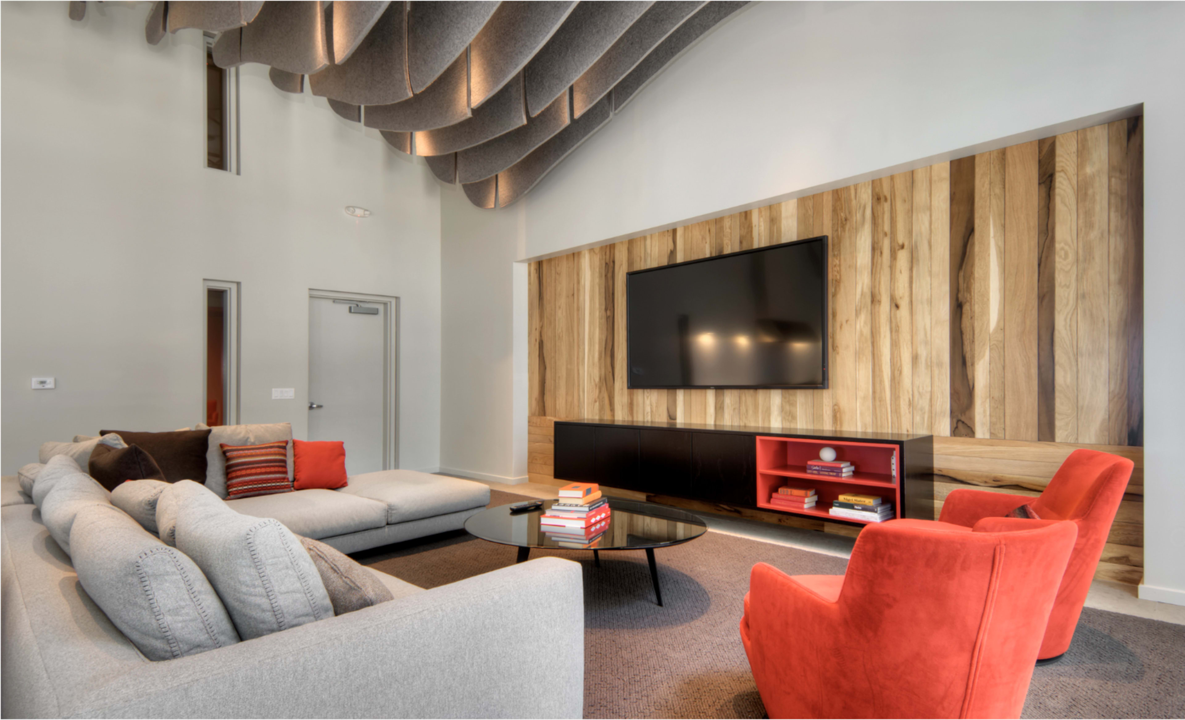 The image shows a modern living room with a large grey sectional sofa, a round glass coffee table, two bright orange chairs, and a feature wall that includes a mounted TV and wooden paneling.