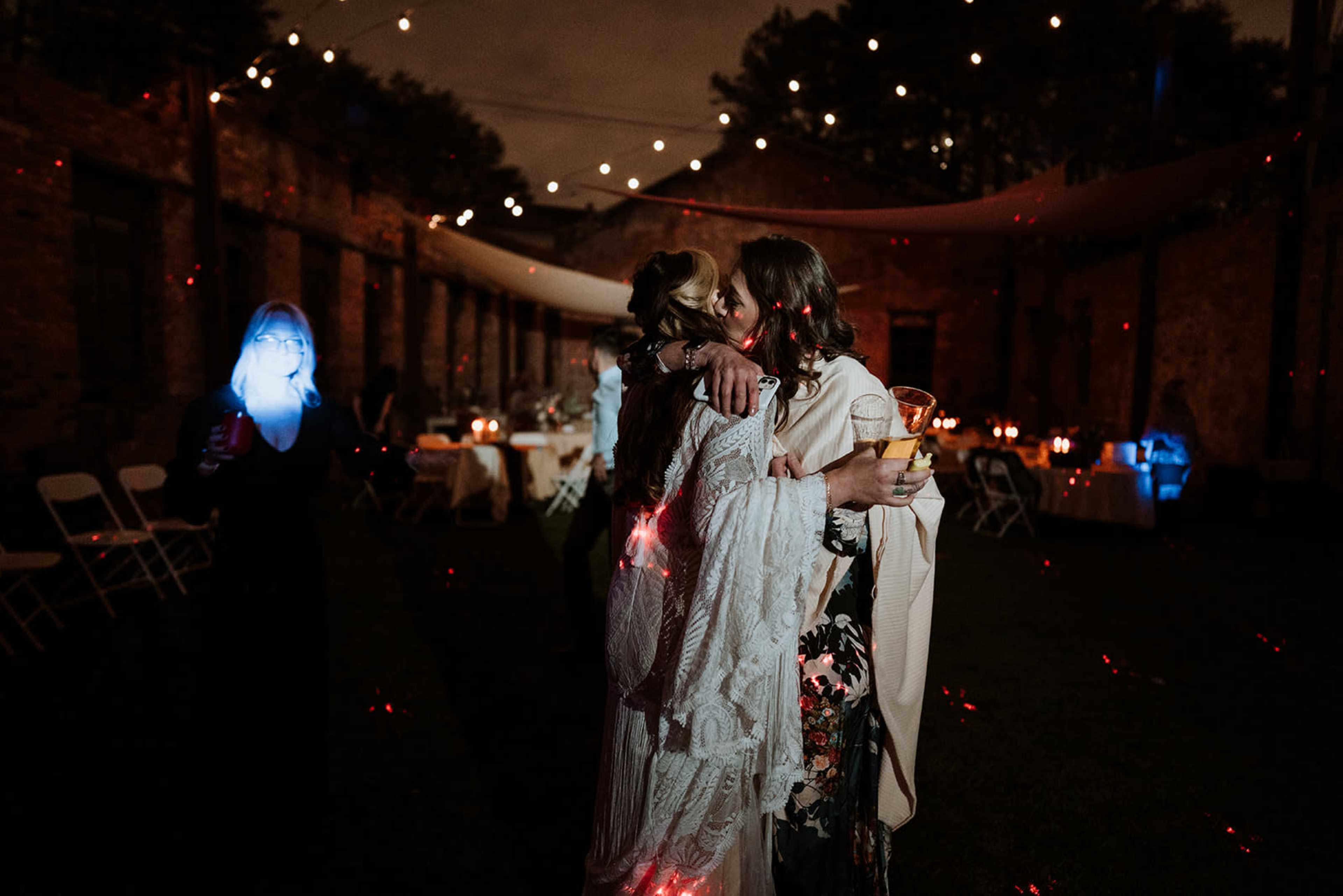Two women embrace while holding drinks in a dimly lit outdoor space adorned with string lights and tables set for a gathering.