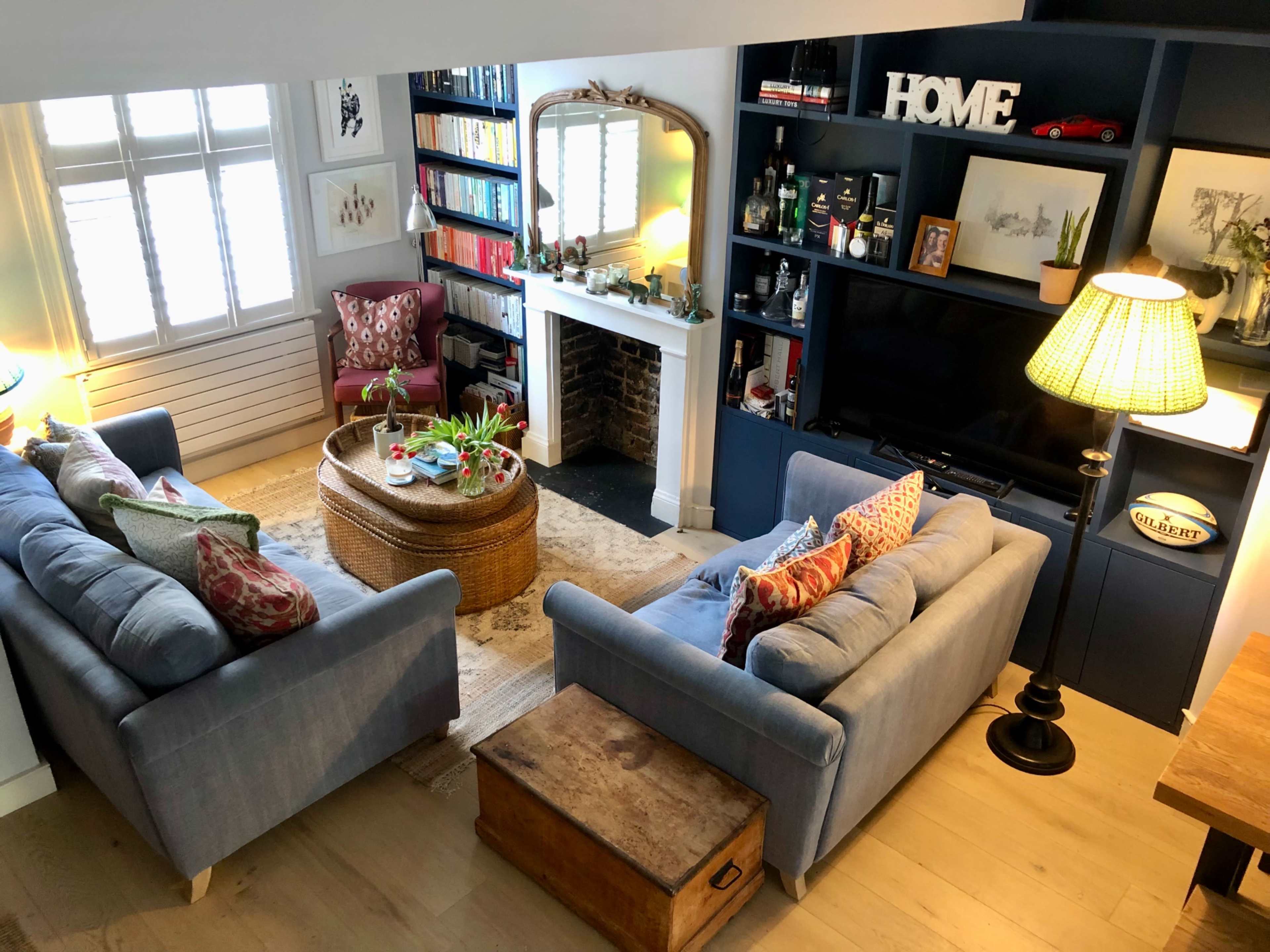 The image shows a cozy living room with two blue sofas facing a wicker coffee table, a fireplace surrounded by shelves, and a wall-mounted television.