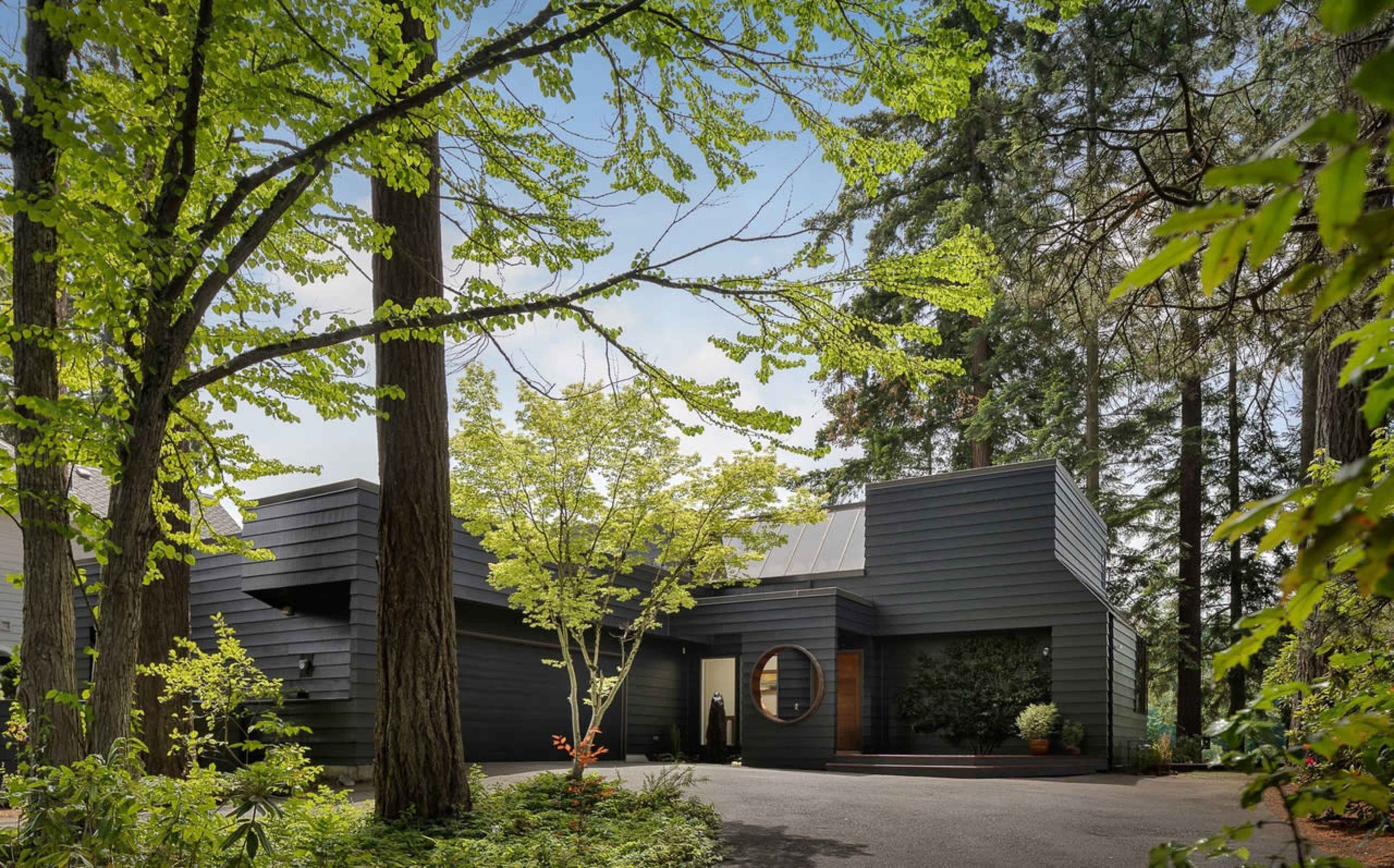 A modern black house with geometric shapes is surrounded by tall trees and lush greenery.