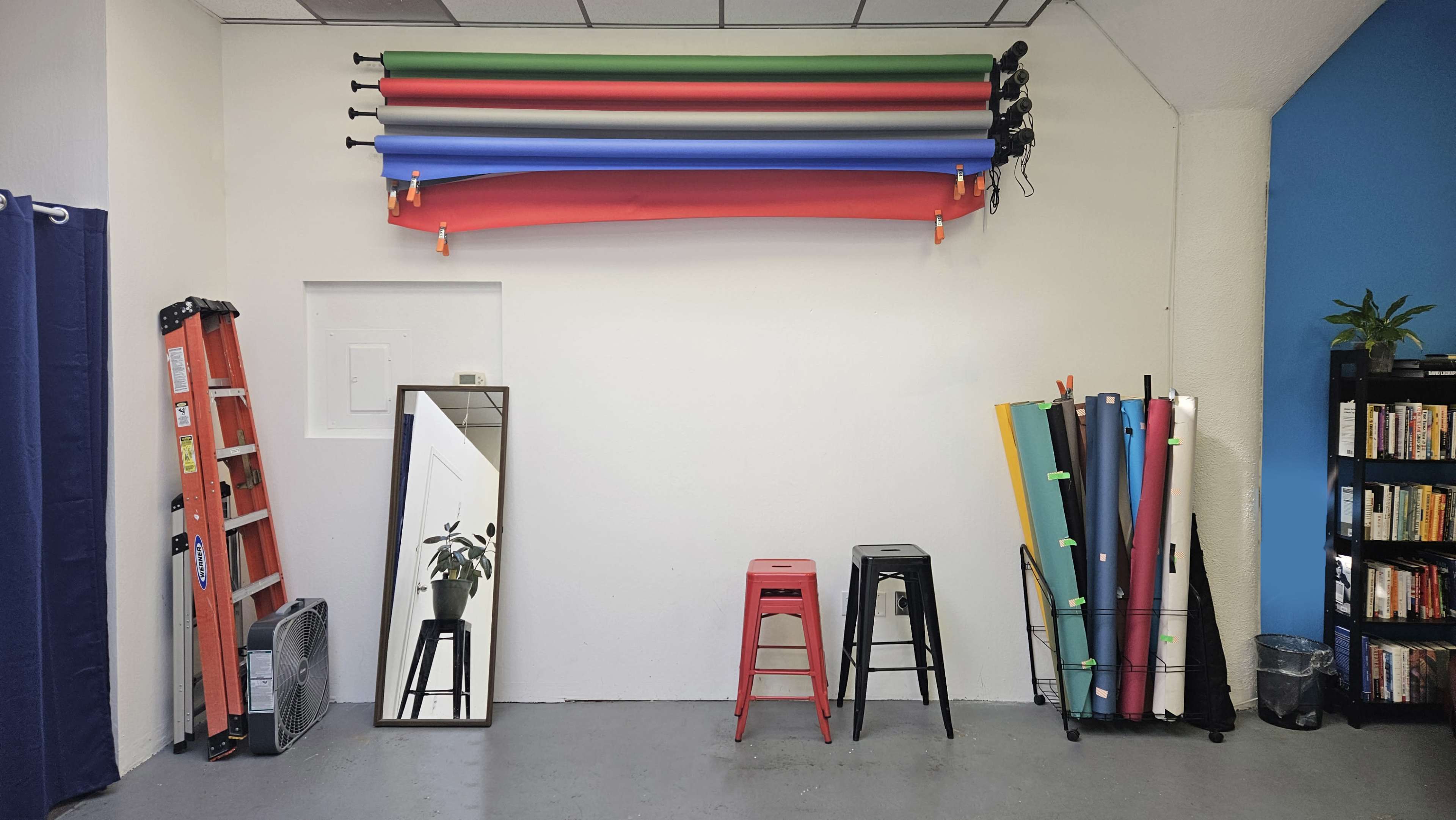 A studio space with a ladder, a mirror, colored backdrops hung on the wall, and several stools arranged beside a bookshelf.