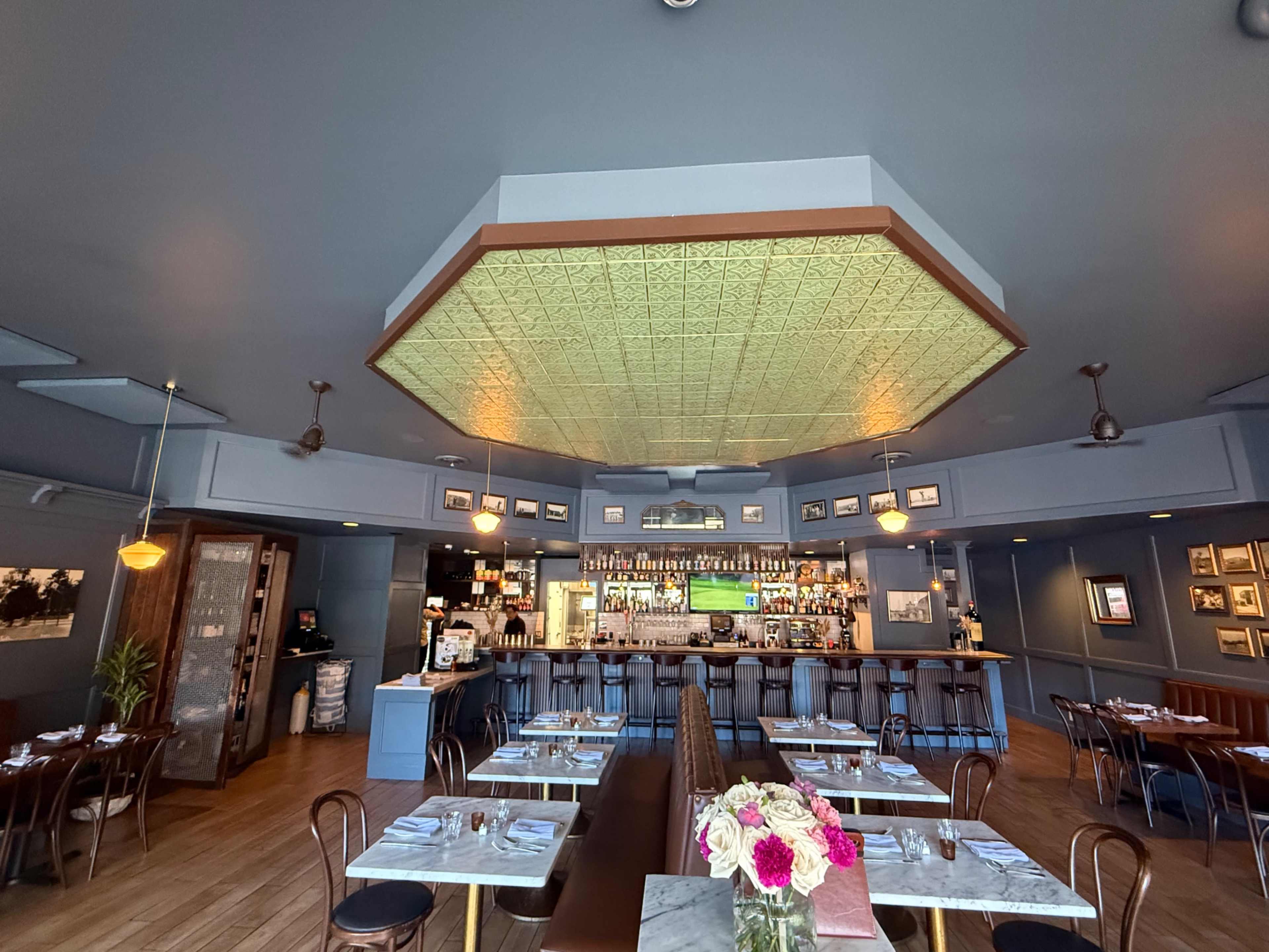 The image shows a restaurant interior with a bar area, featuring a geometric patterned ceiling, wooden flooring, and tables set for dining.