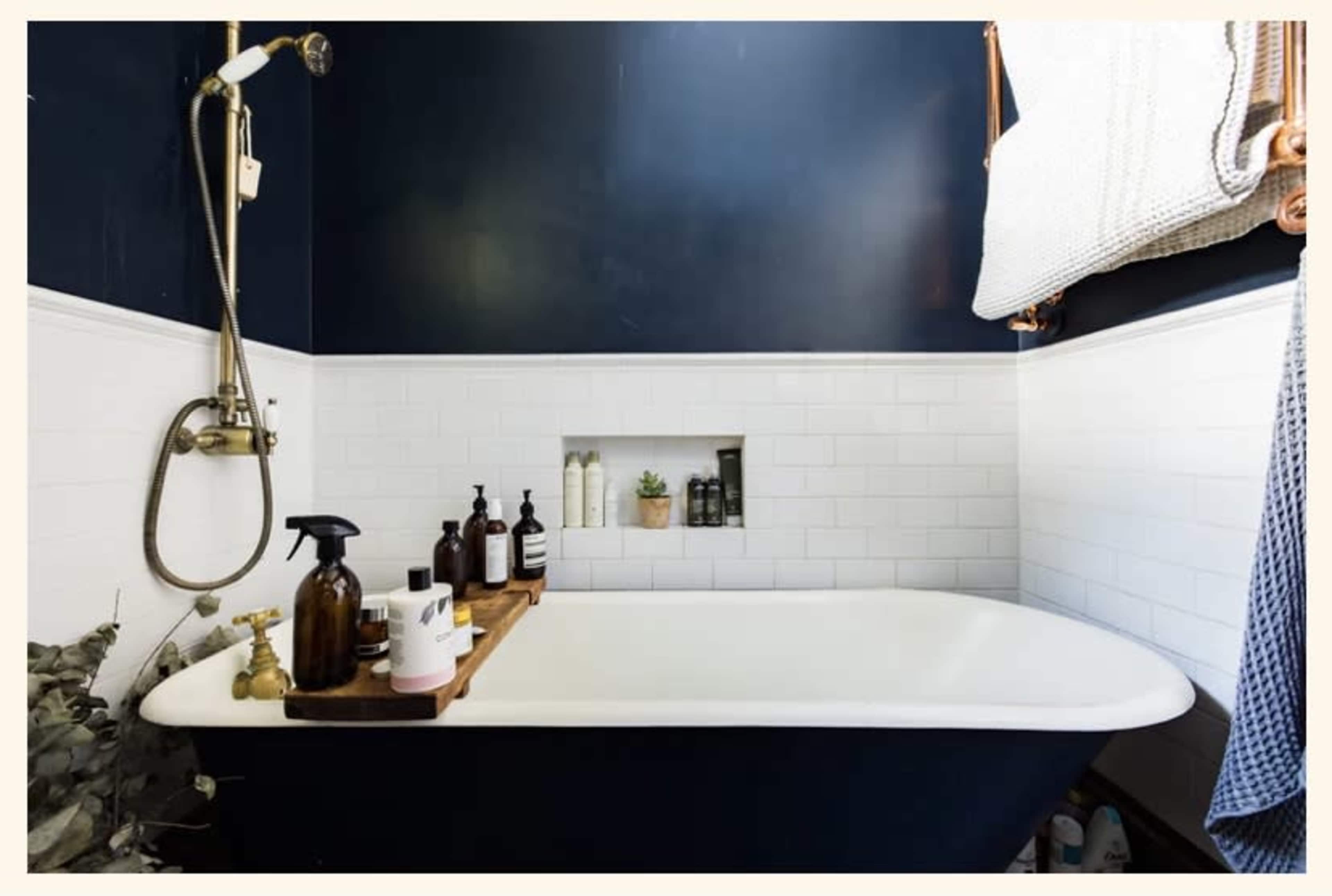 A bathtub in a bathroom with dark blue walls, white tile wainscoting, and a shelf with various grooming products and a small plant.