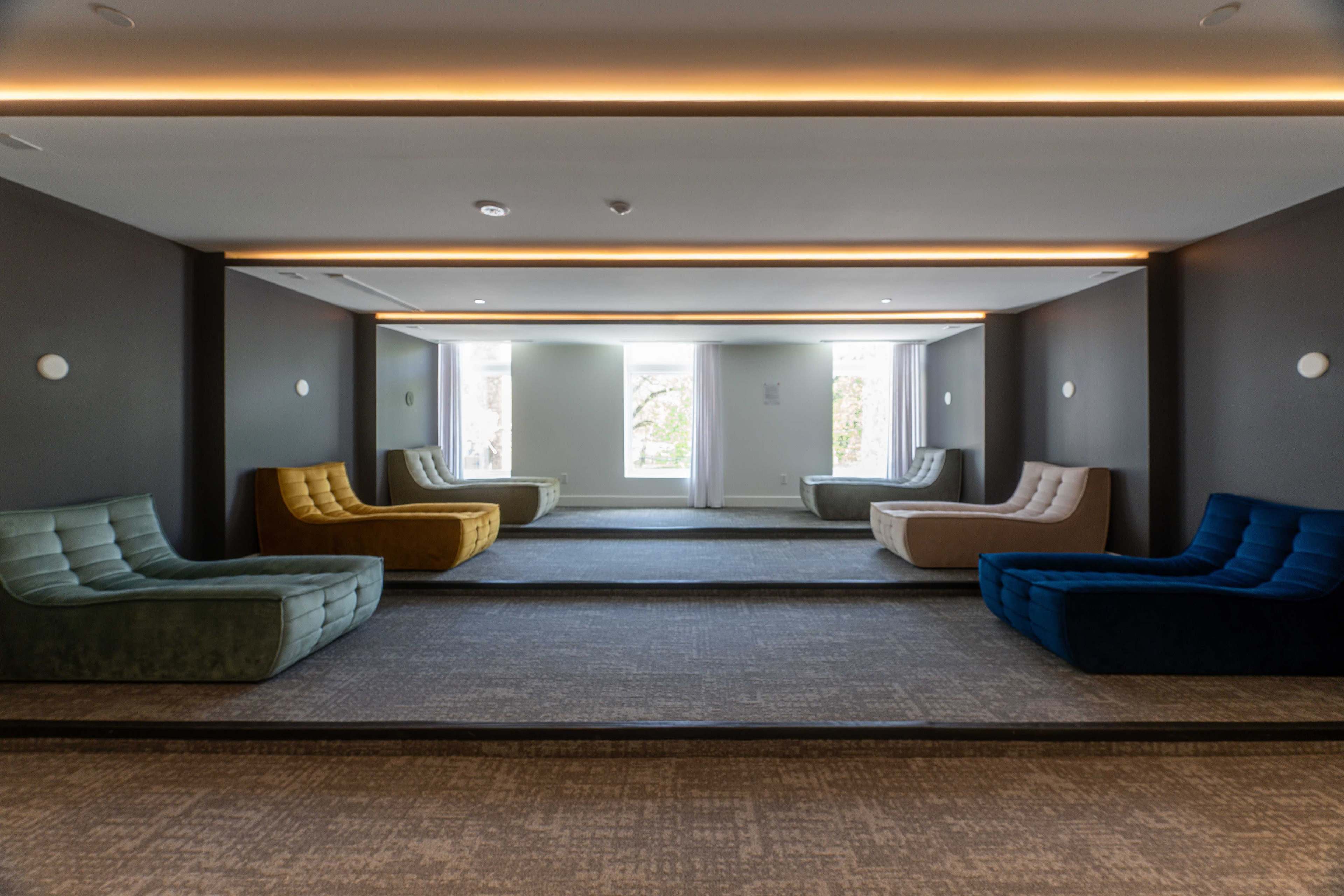 The image shows a modern lounge area featuring several chaise lounges in various colors arranged symmetrically around a central space, with large windows allowing natural light to enter.