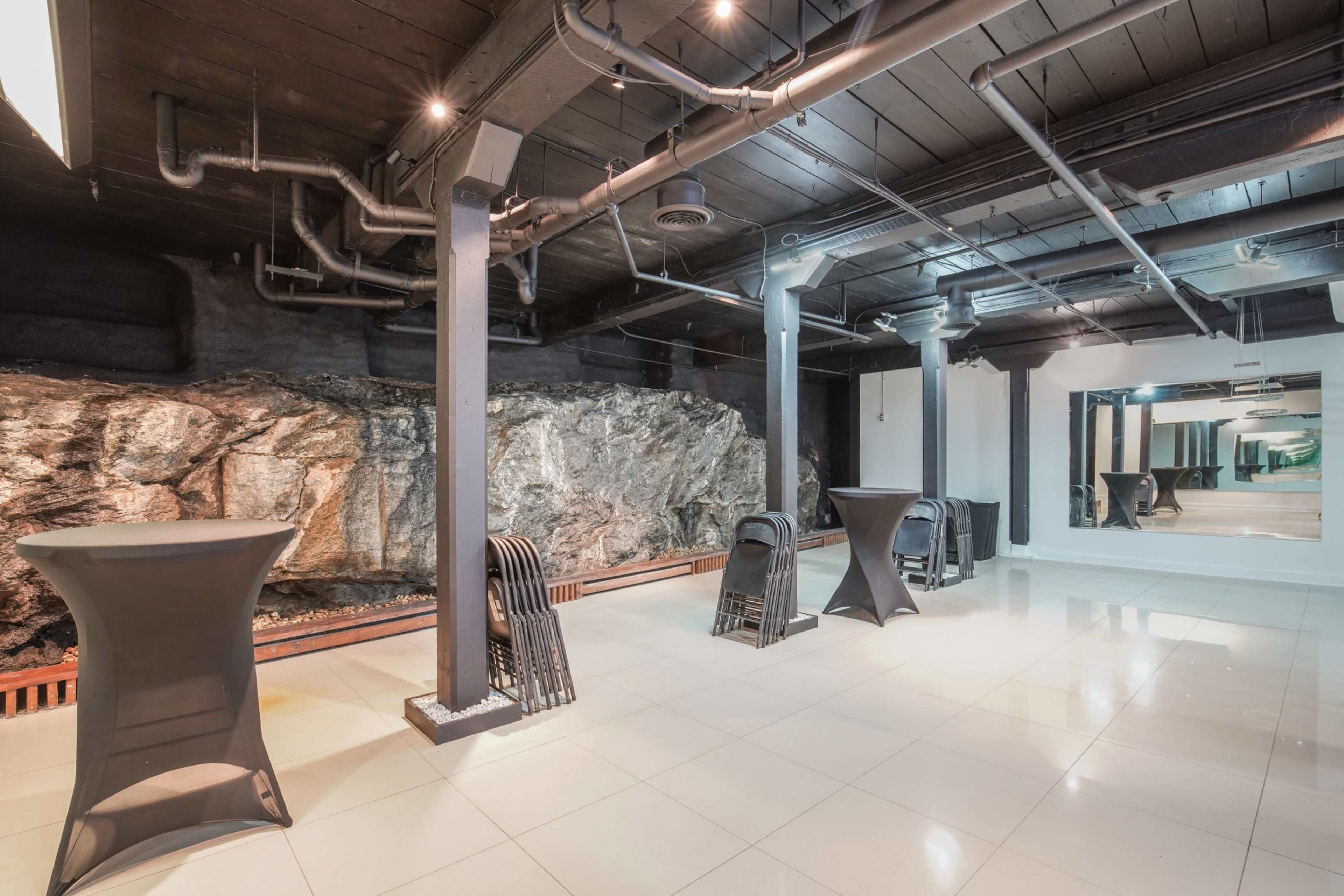 The image shows an industrial-style indoor space featuring a large rock wall, black columns, and several black cocktail tables set on a tiled floor.