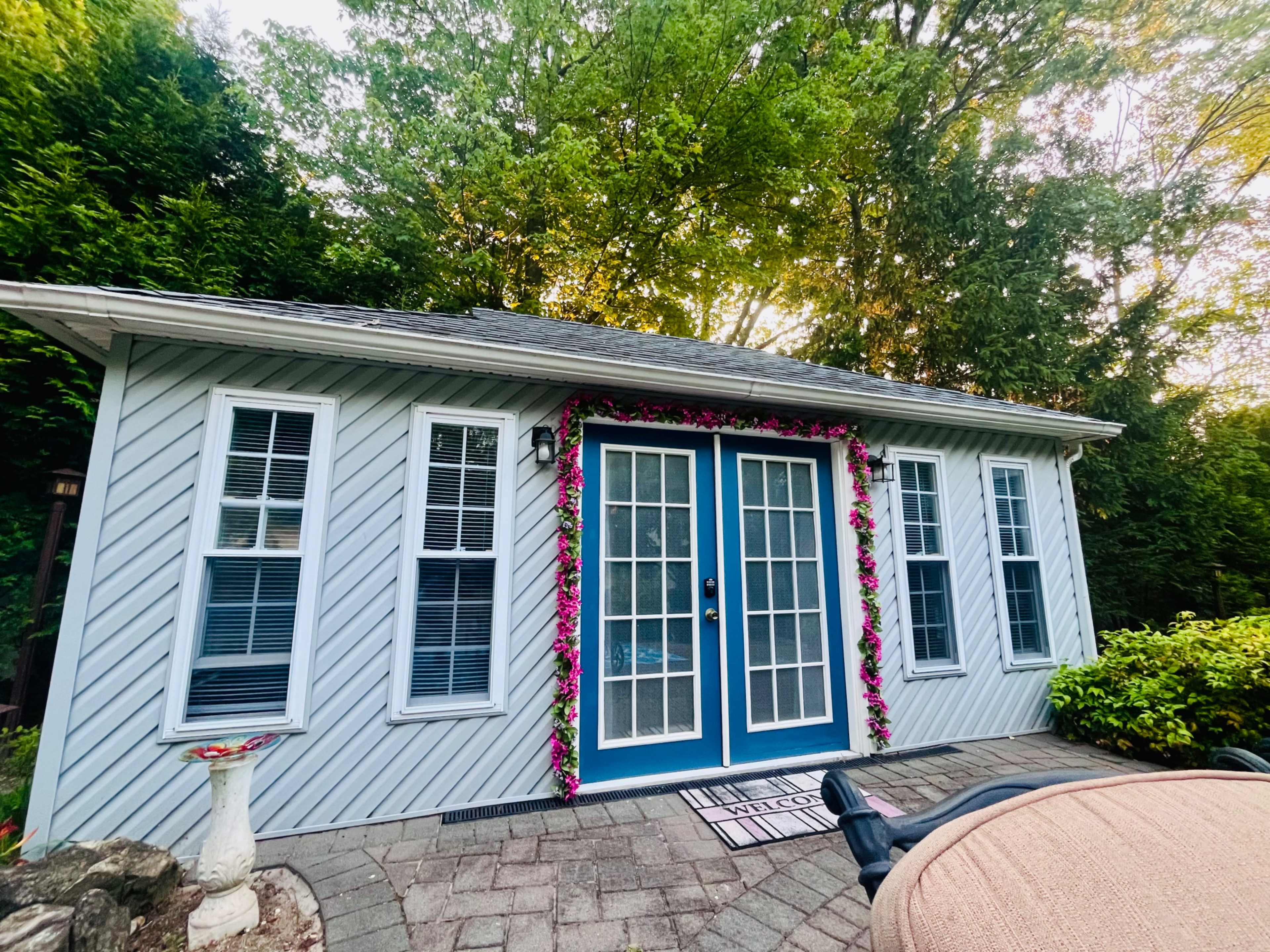 A small house with blue double doors and flower decorations is surrounded by trees and a paved area.