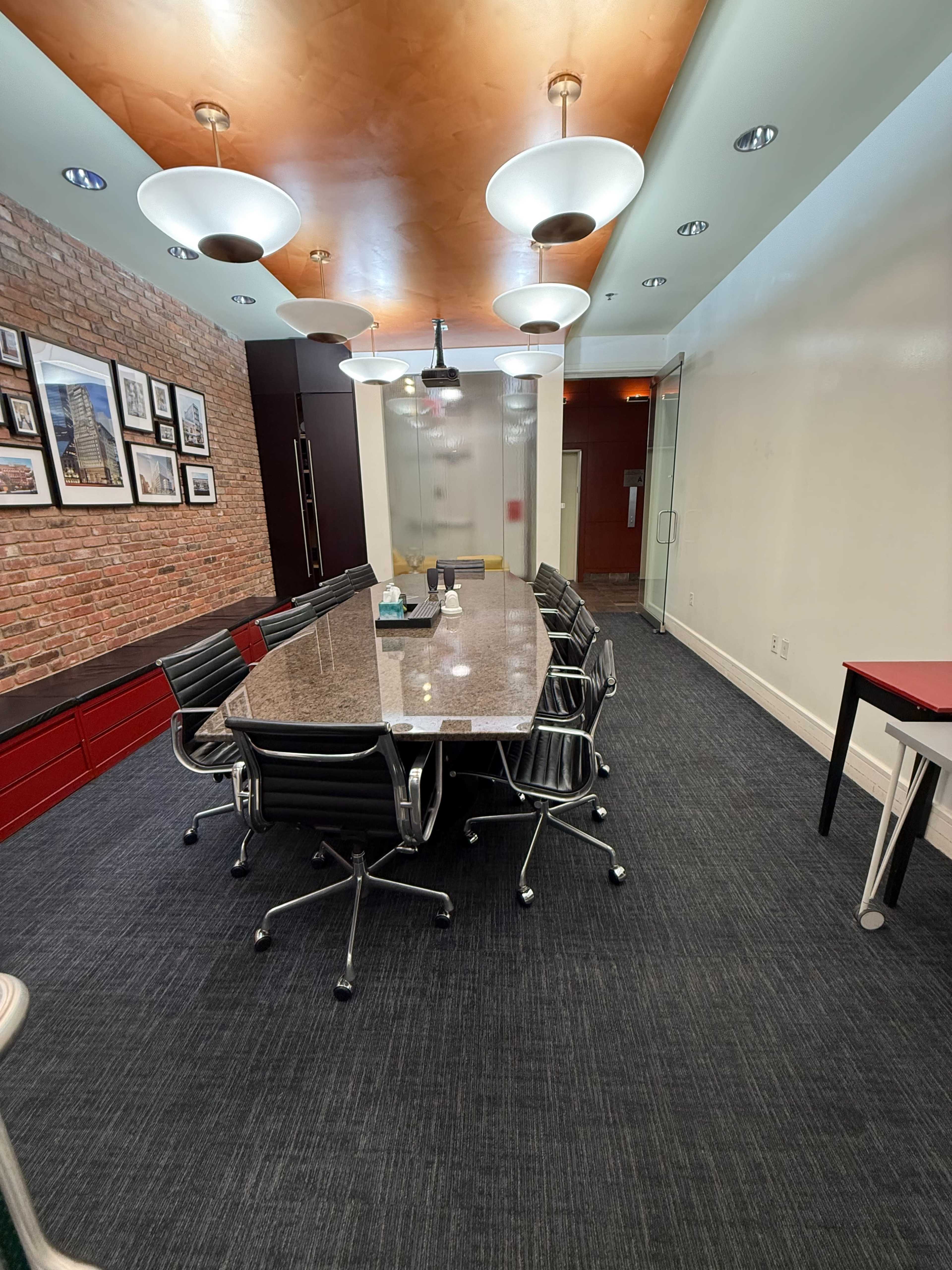 A modern conference room features a long granite table surrounded by ergonomic black chairs, with exposed brick walls and unique ceiling lights.