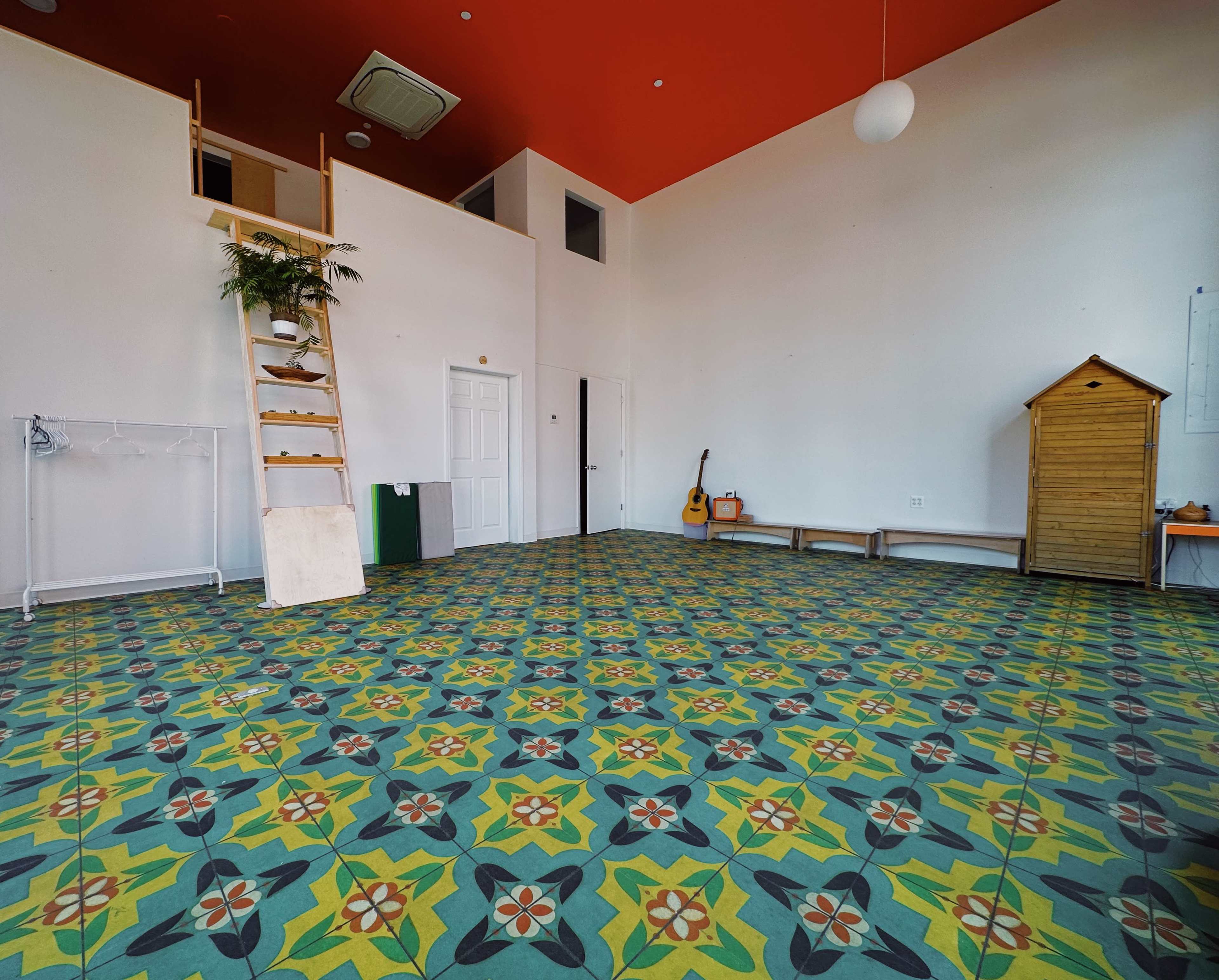 A spacious, brightly colored room with patterned flooring, a small wooden shed, a guitar resting against the wall, and a ladder leading to a second level.