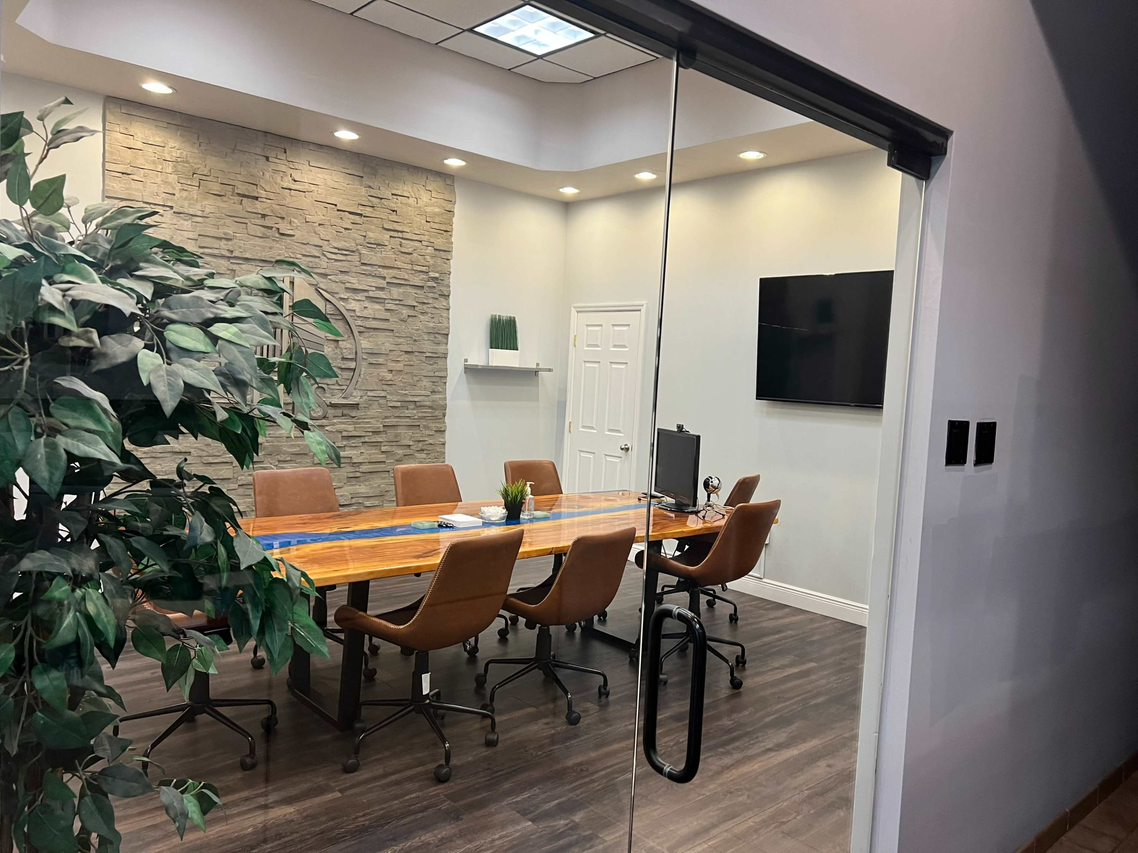 A modern conference room with a long wooden table surrounded by brown chairs, and a stone accent wall, visible through a glass door.