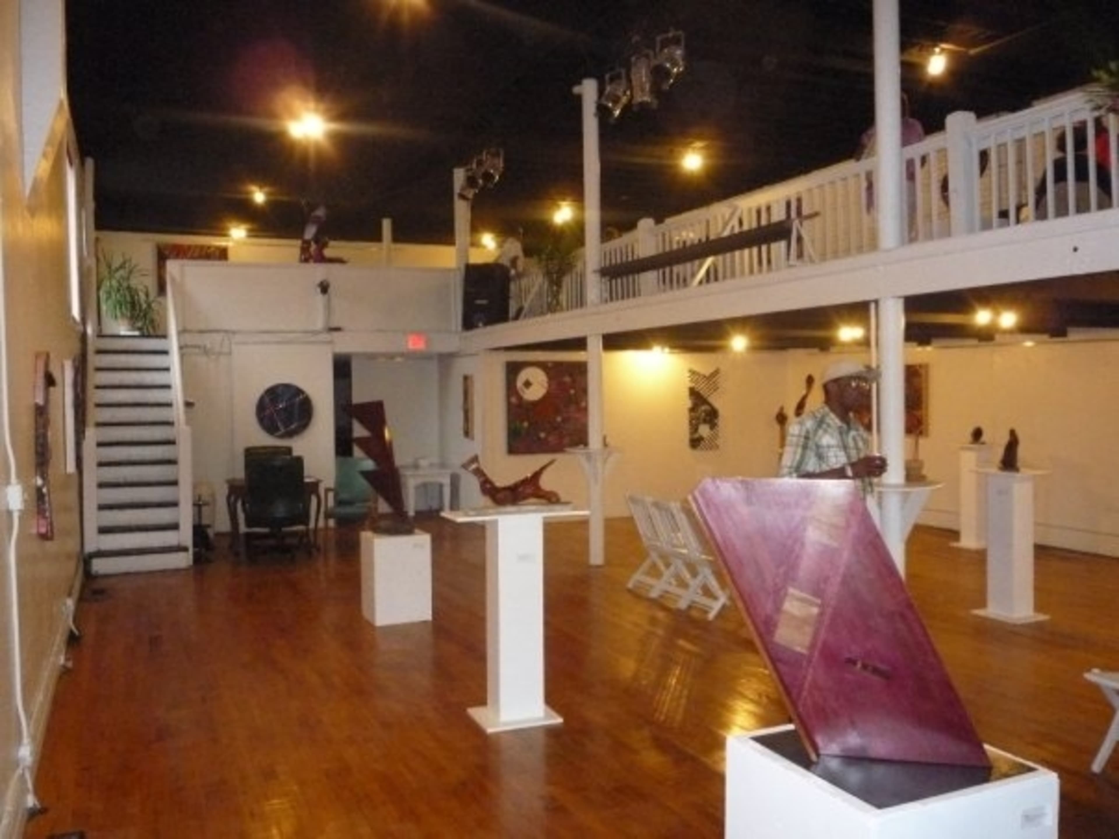 An art gallery featuring wooden sculptures displayed on pedestals, with wooden flooring and a staircase leading to a balcony above.