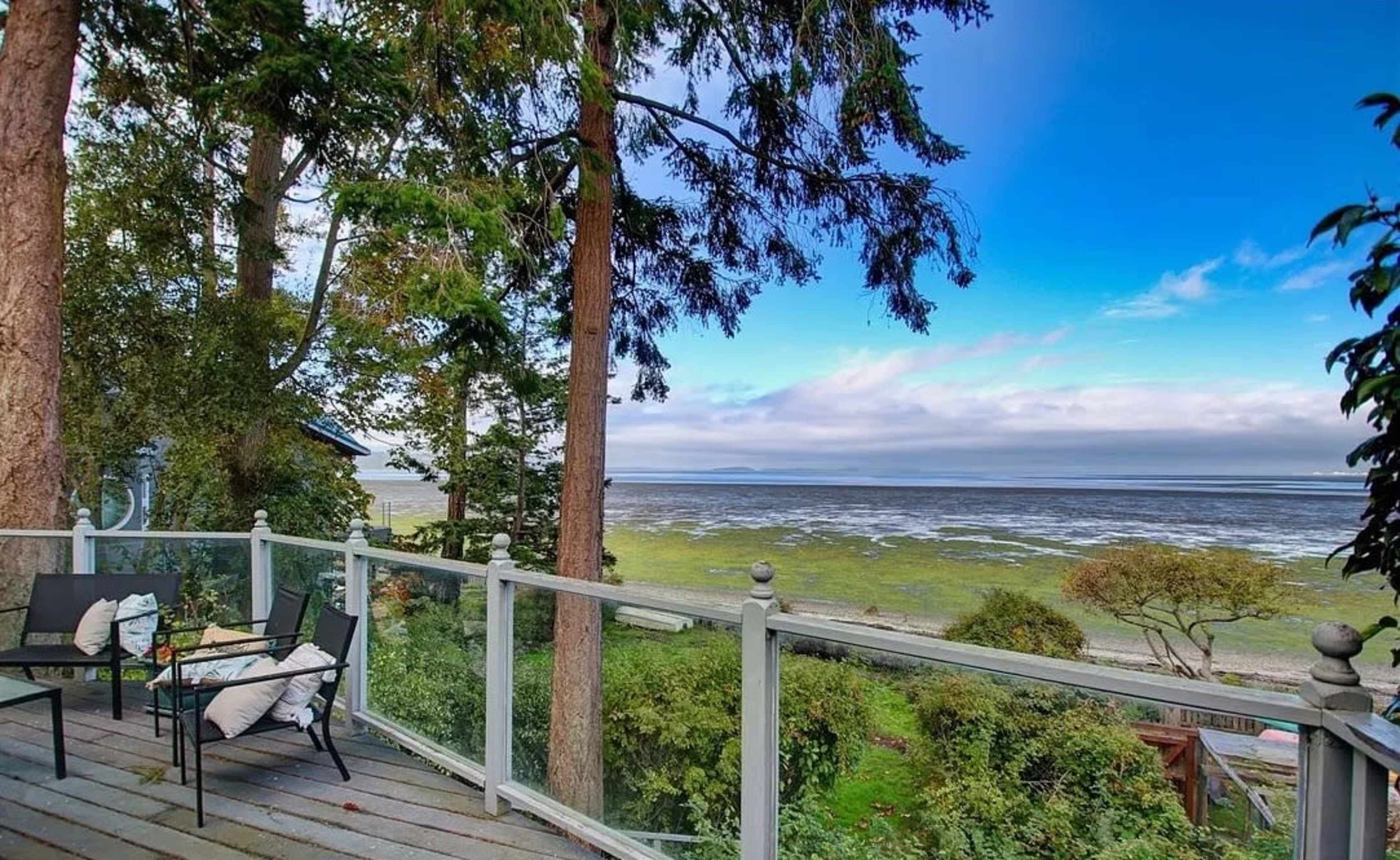 A wooden deck with chairs overlooks a tidal bay surrounded by trees and low vegetation under a partly cloudy sky.