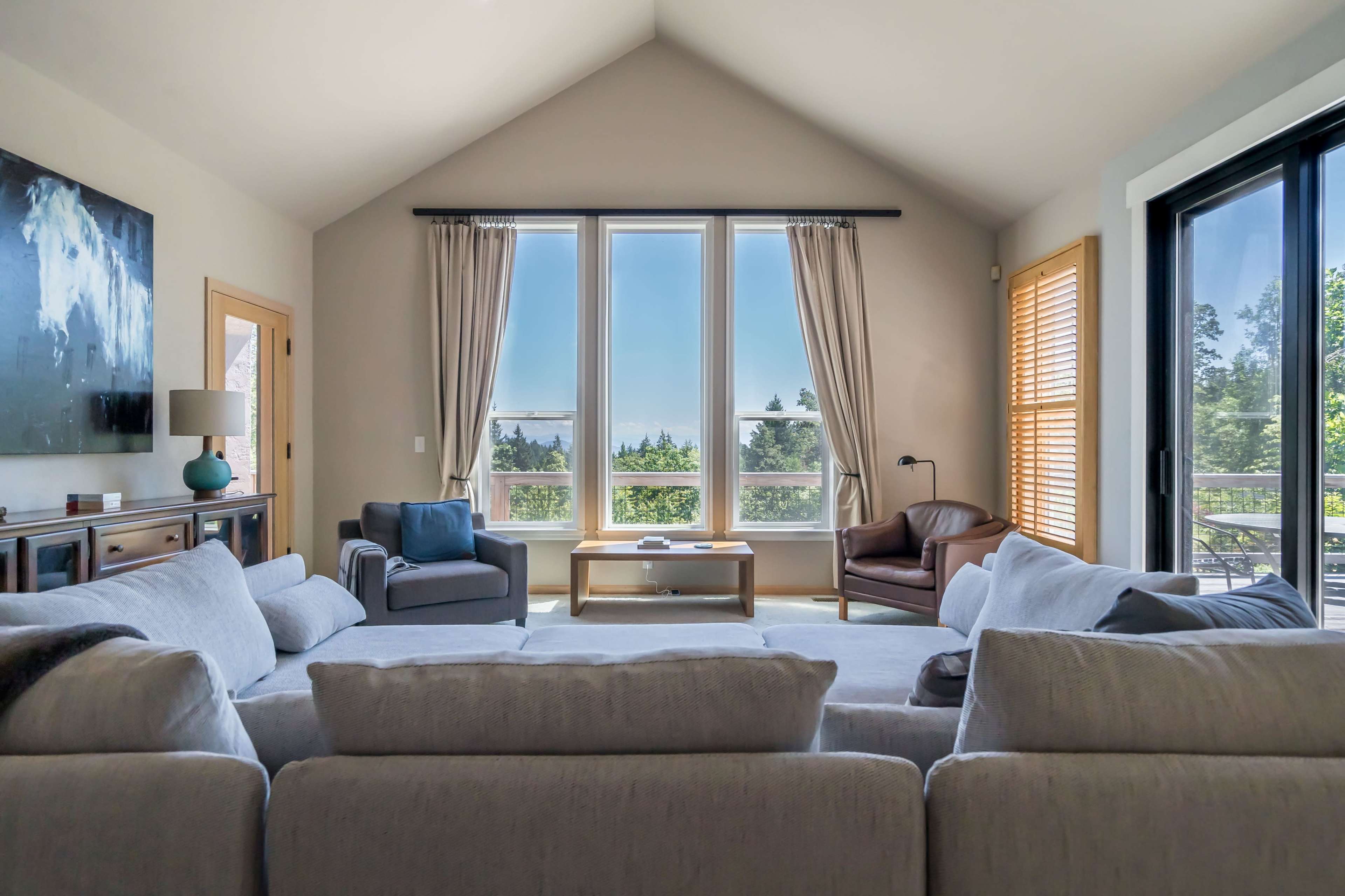 A spacious living room features large windows overlooking a balcony and trees, with a light-colored sofa, armchairs, and a coffee table in the center.