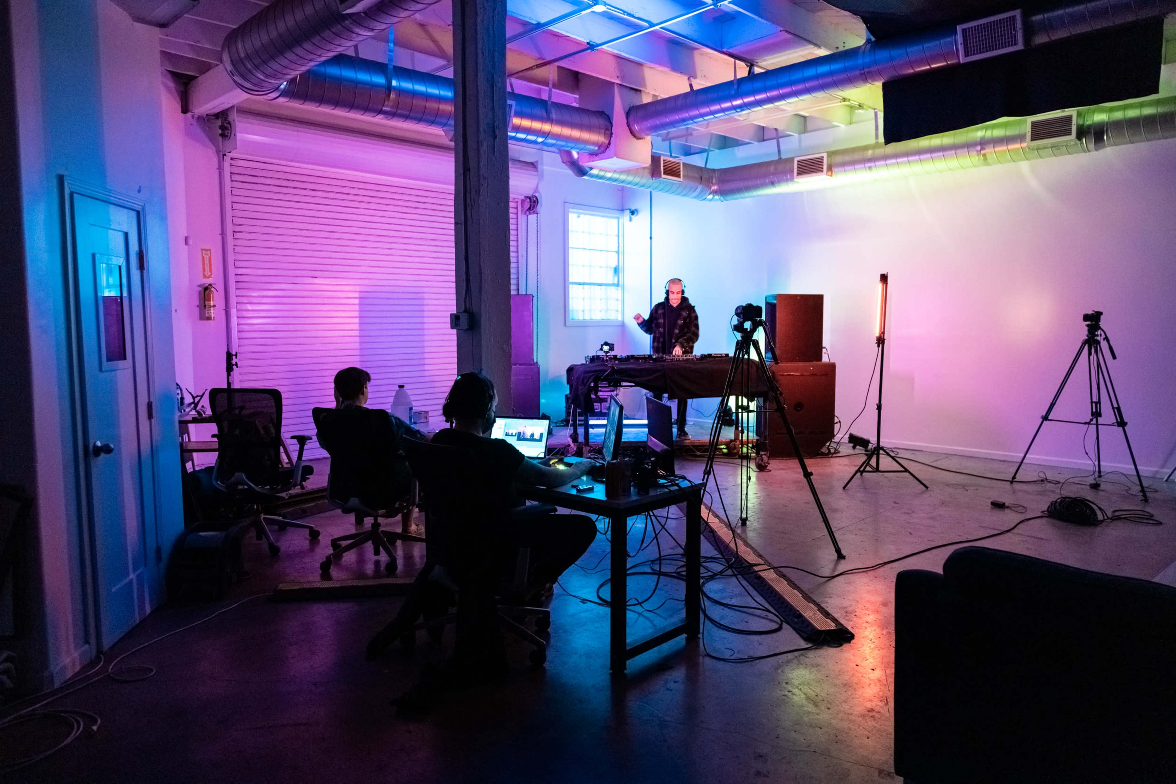 A man stands behind a table with equipment while two individuals sit at a desk with computers in a brightly lit studio space.
