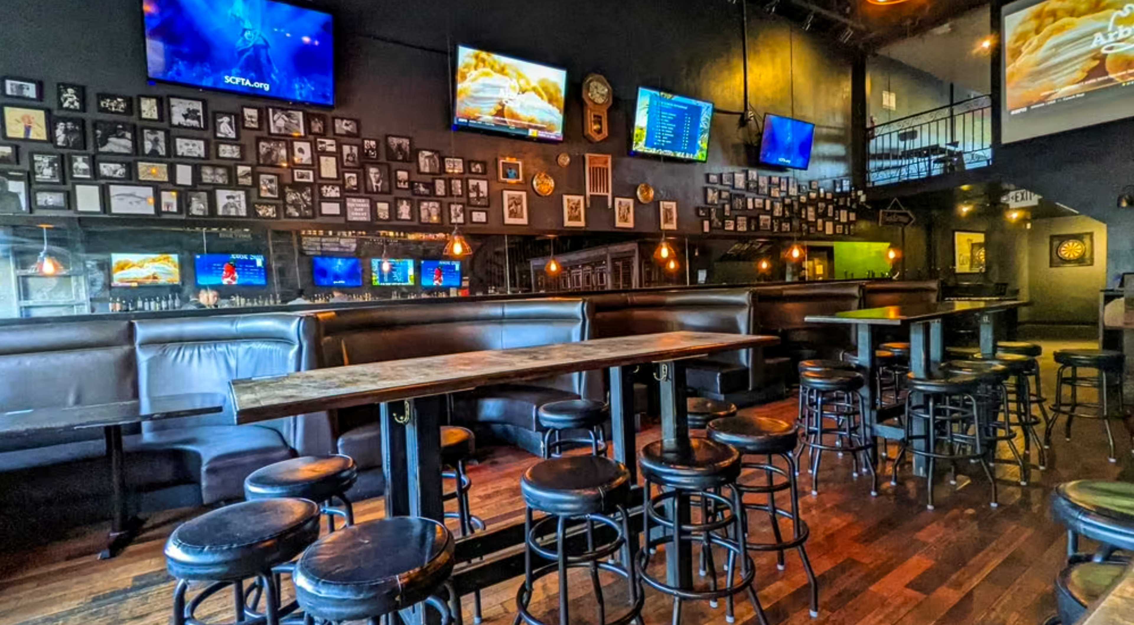 The image shows a dimly lit bar with black leather seating, wooden tables, and walls adorned with framed photos and multiple televisions displaying sports.