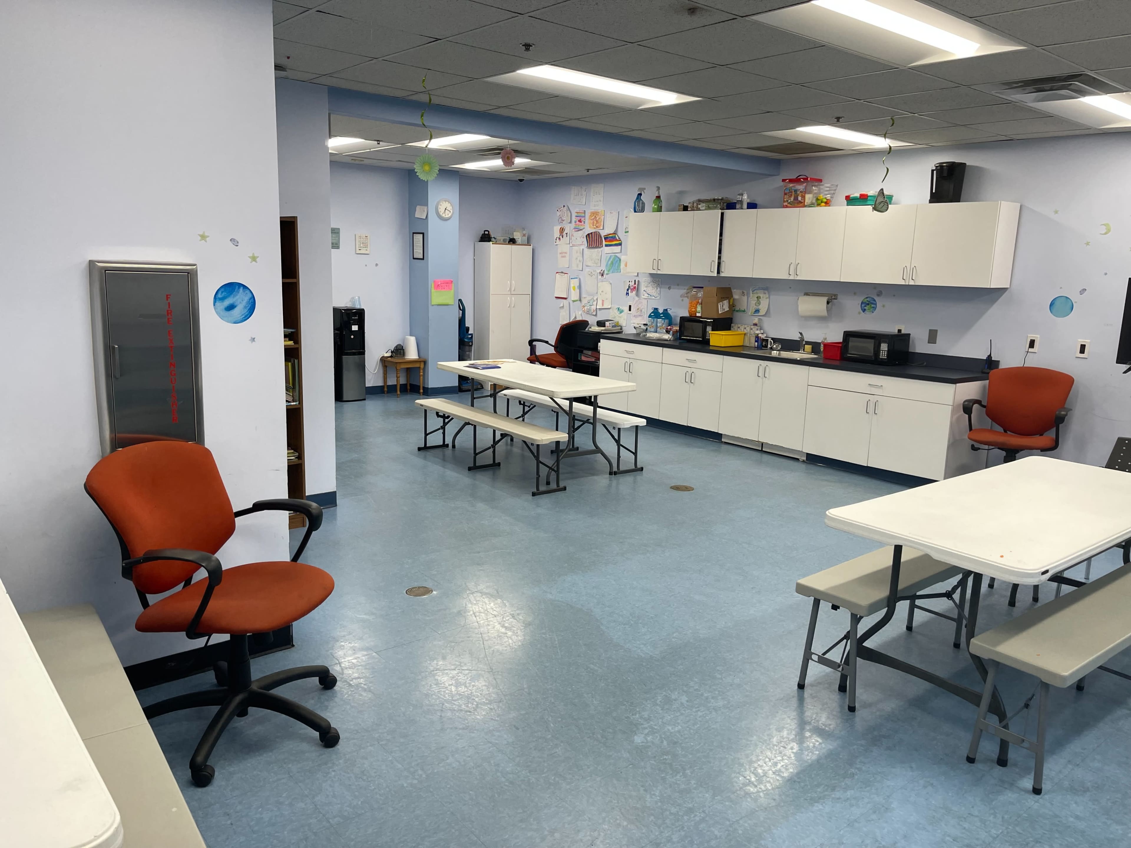A kitchen or break room with several tables and chairs arranged around the space, and cabinets and appliances along the walls.