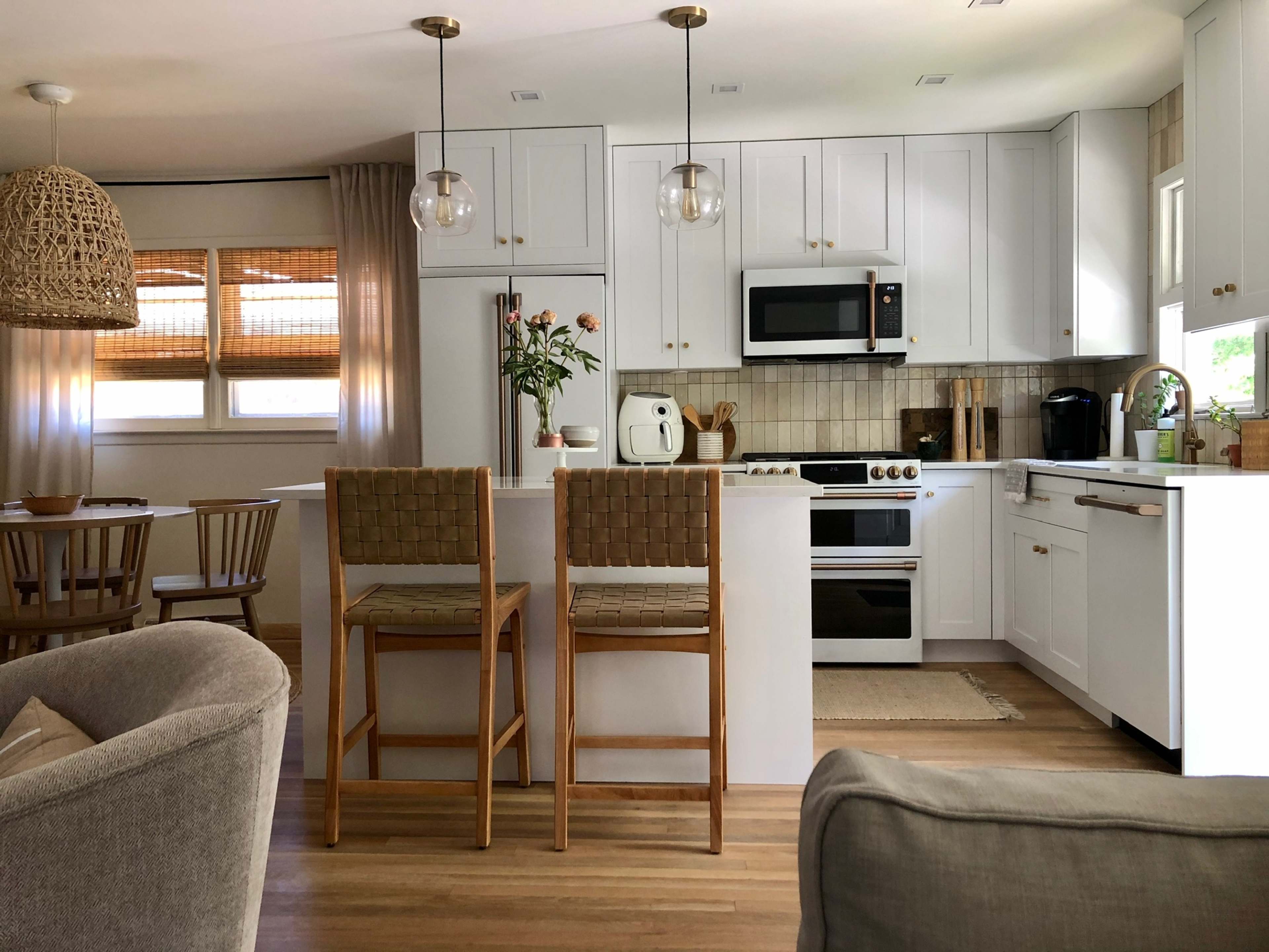 A modern kitchen featuring white cabinets, stainless steel appliances, and two barstools at a small island, with a dining area visible in the background.