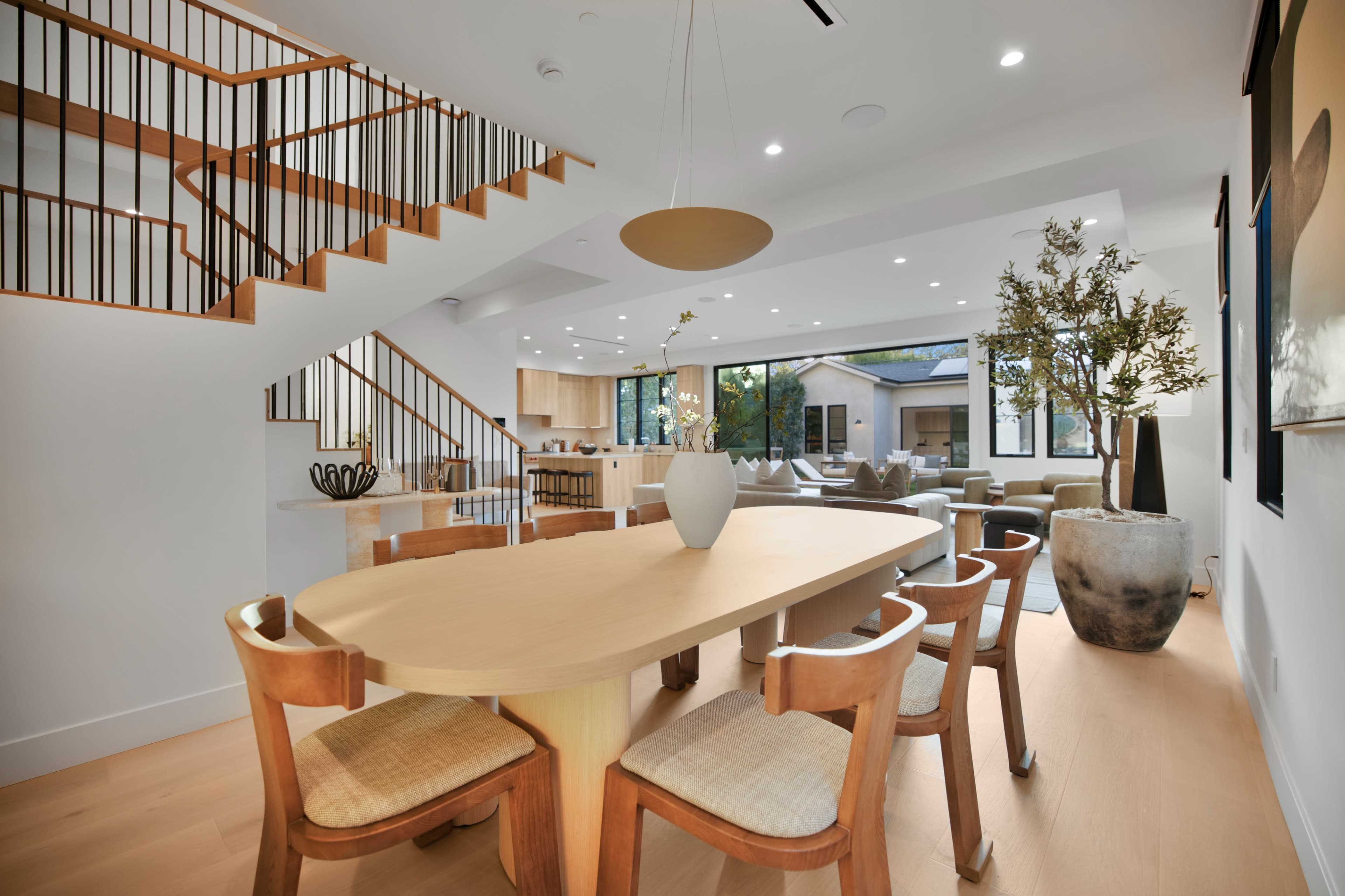 The image shows a modern dining area featuring a round table with wooden chairs, a staircase in the background, and large windows that open to an outdoor view.