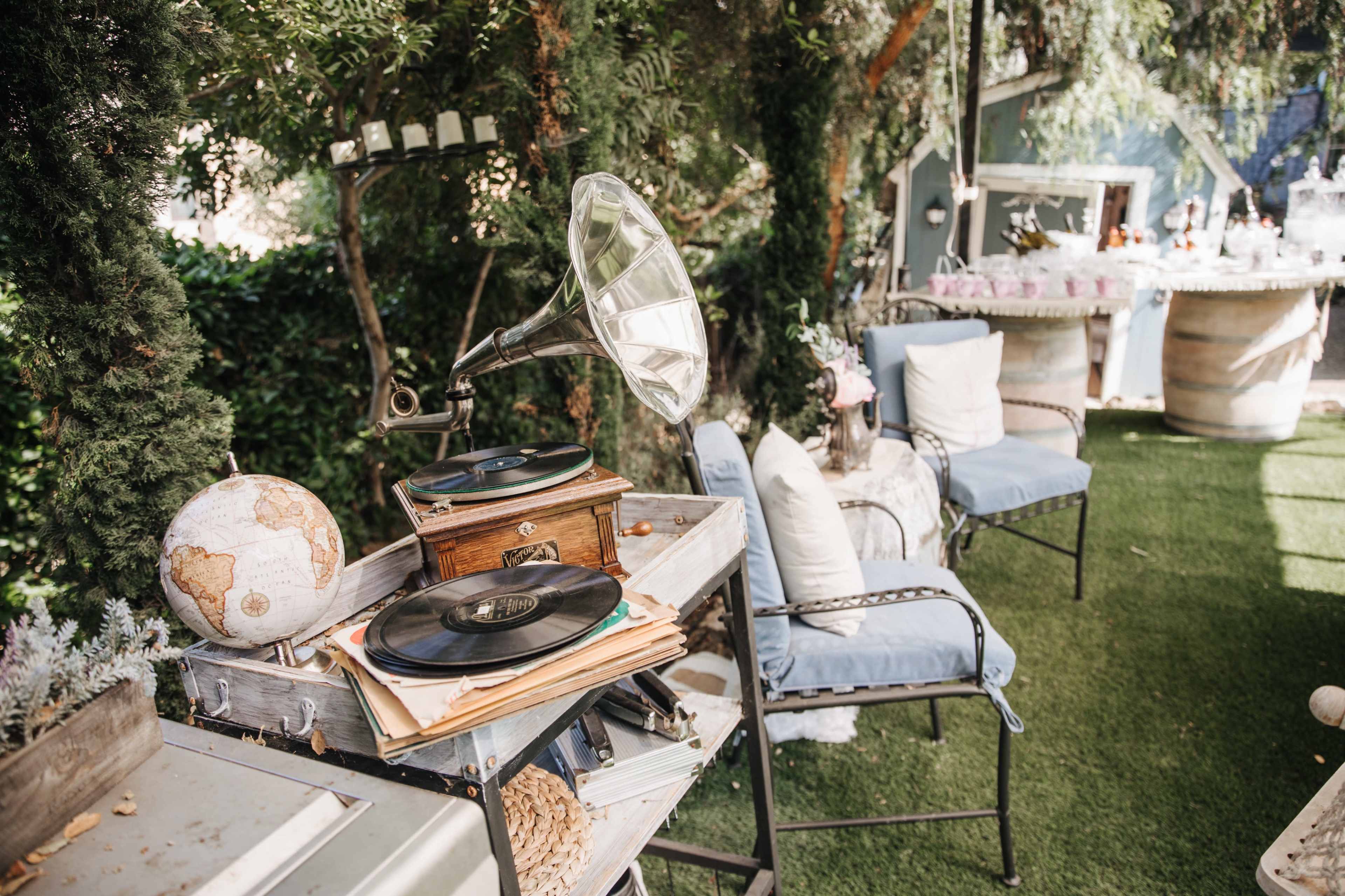 A vintage gramophone with records is placed on a wooden table next to a decorative globe in a garden setting.