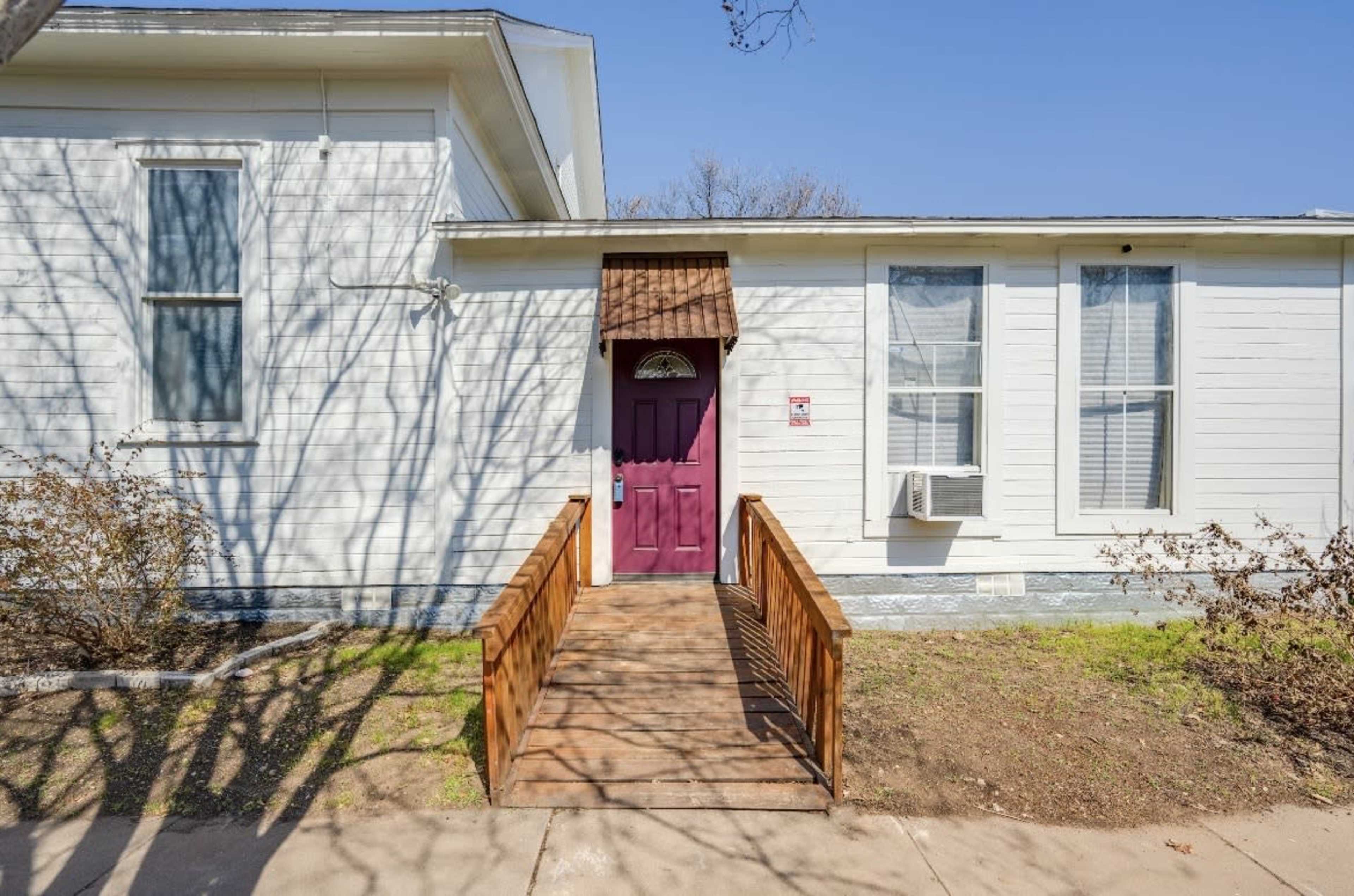 The image shows a front view of a house with a purple door and a wooden ramp leading up to it, flanked by two windows.