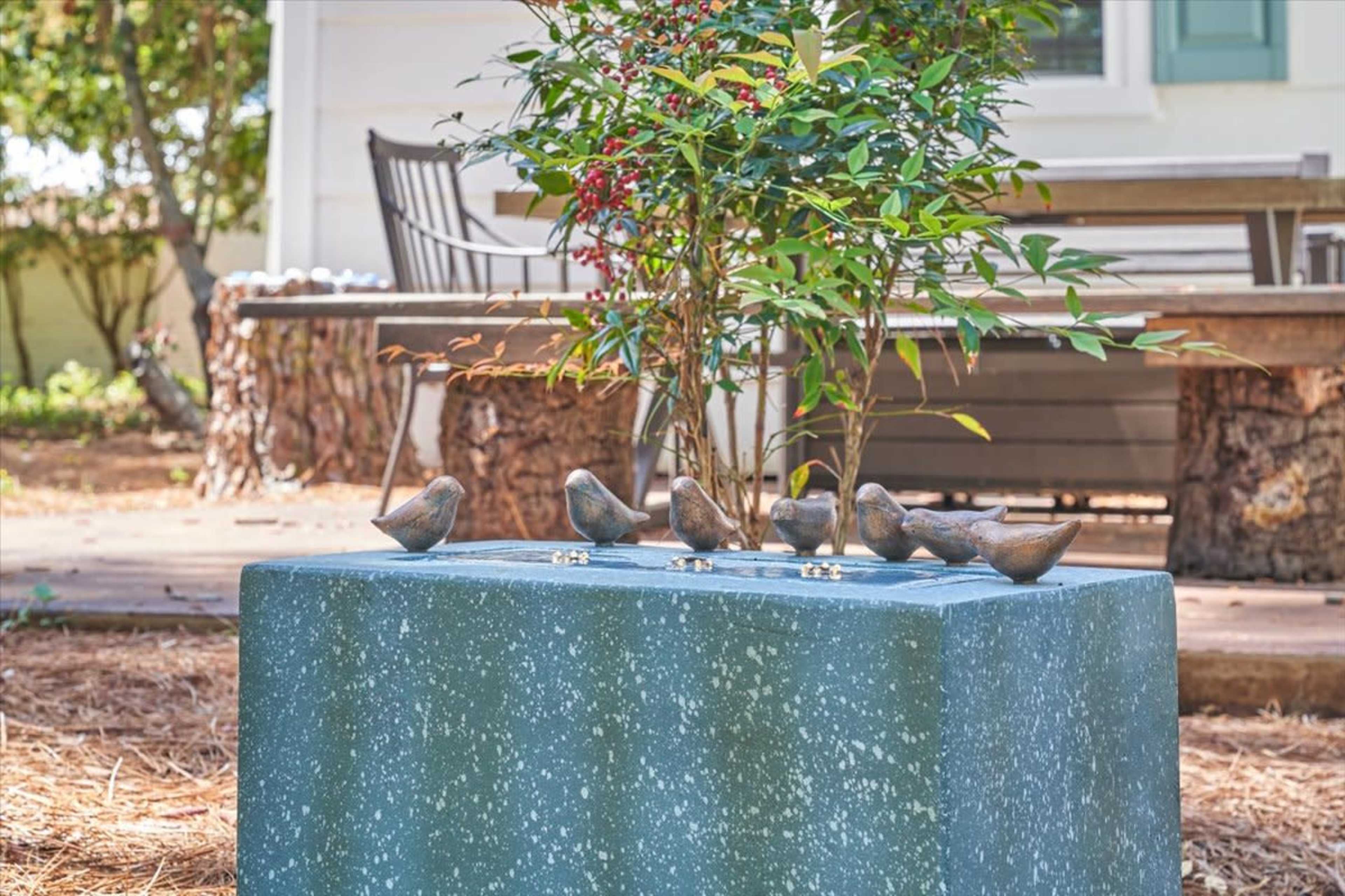A stone pedestal with five bronze bird sculptures is surrounded by green foliage and backed by a wooden bench.