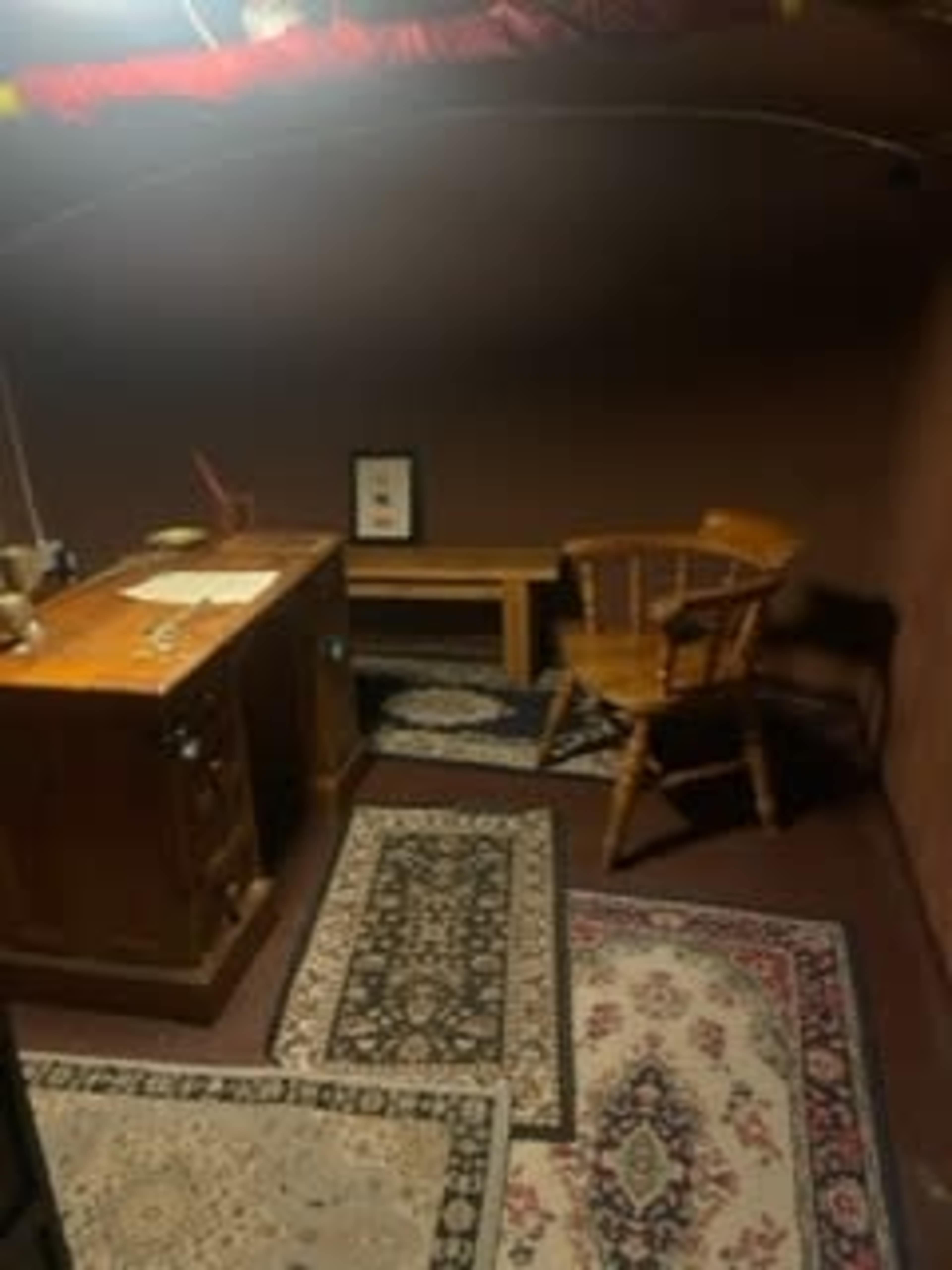 The image shows a sparsely furnished room with a wooden desk, a chair, and rugs on the floor.