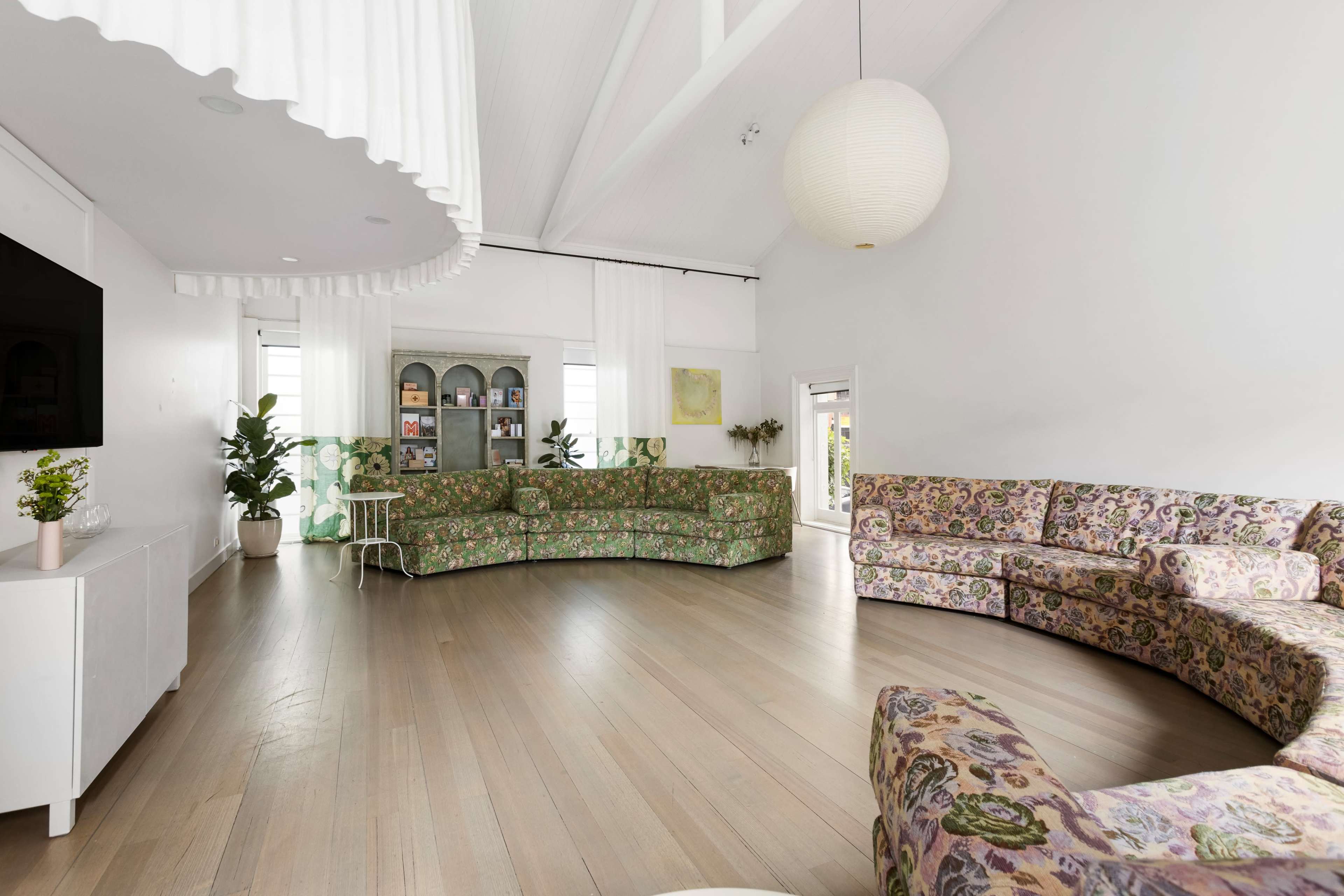 The room features a large, curved sofa with a floral pattern, wooden flooring, and large windows allowing natural light to enter.