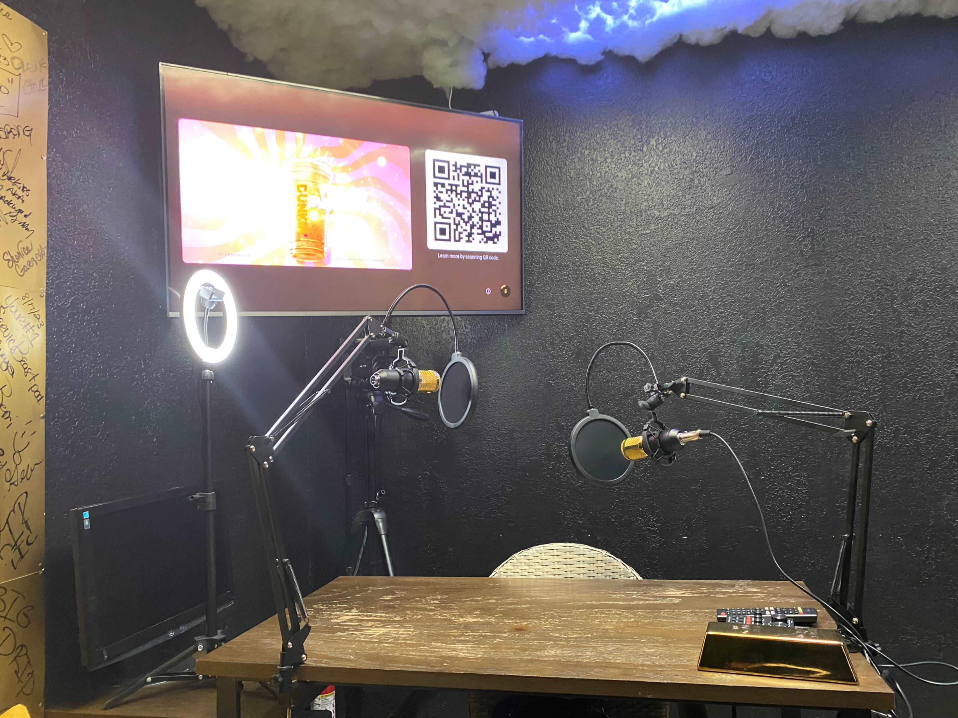 The image shows a small recording studio with two microphone setups, a wooden table, and a screen displaying a visual element on the wall.