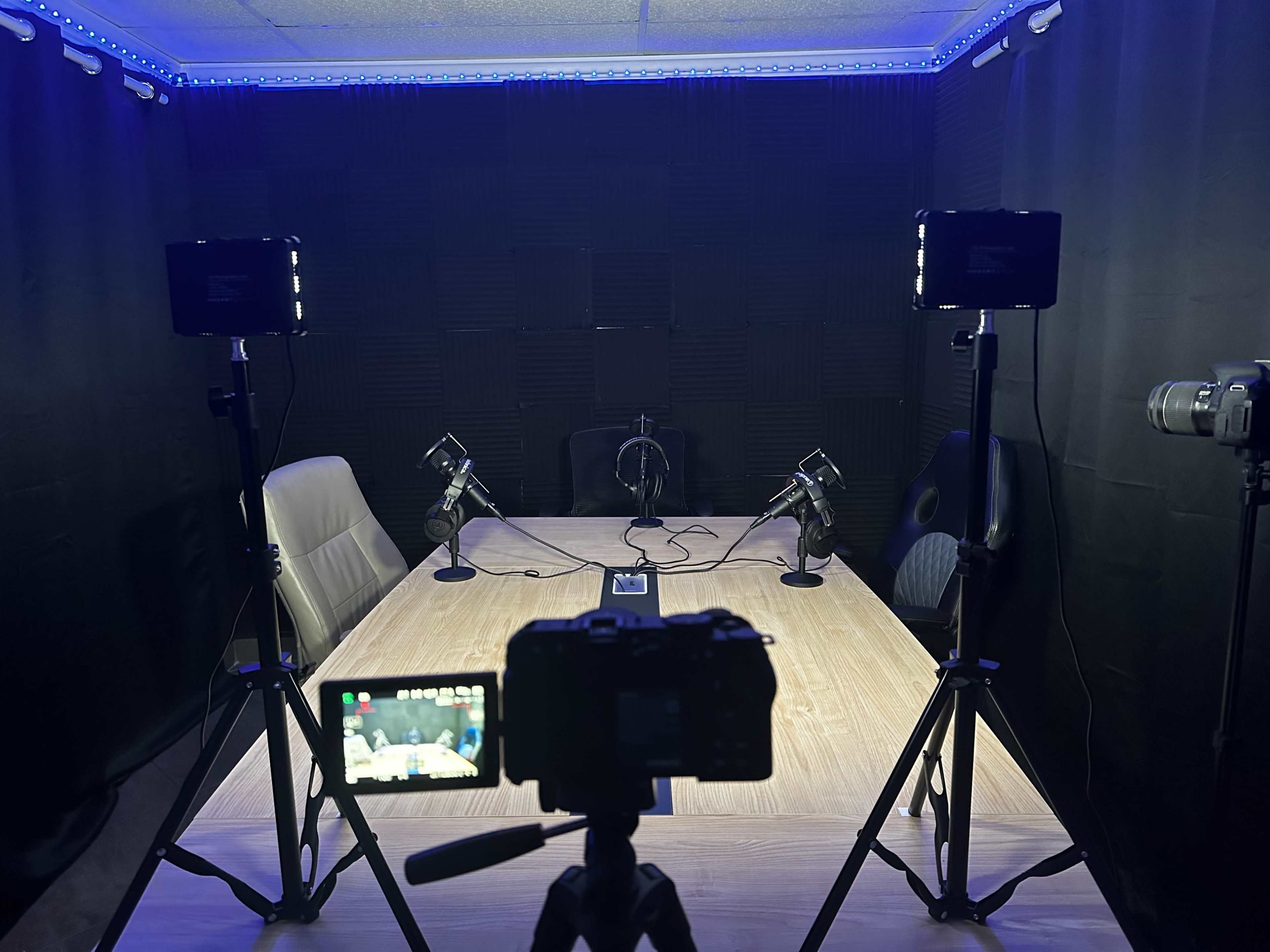 A dimly lit recording studio features a large table with microphones, cameras on tripods, and soundproofed walls.