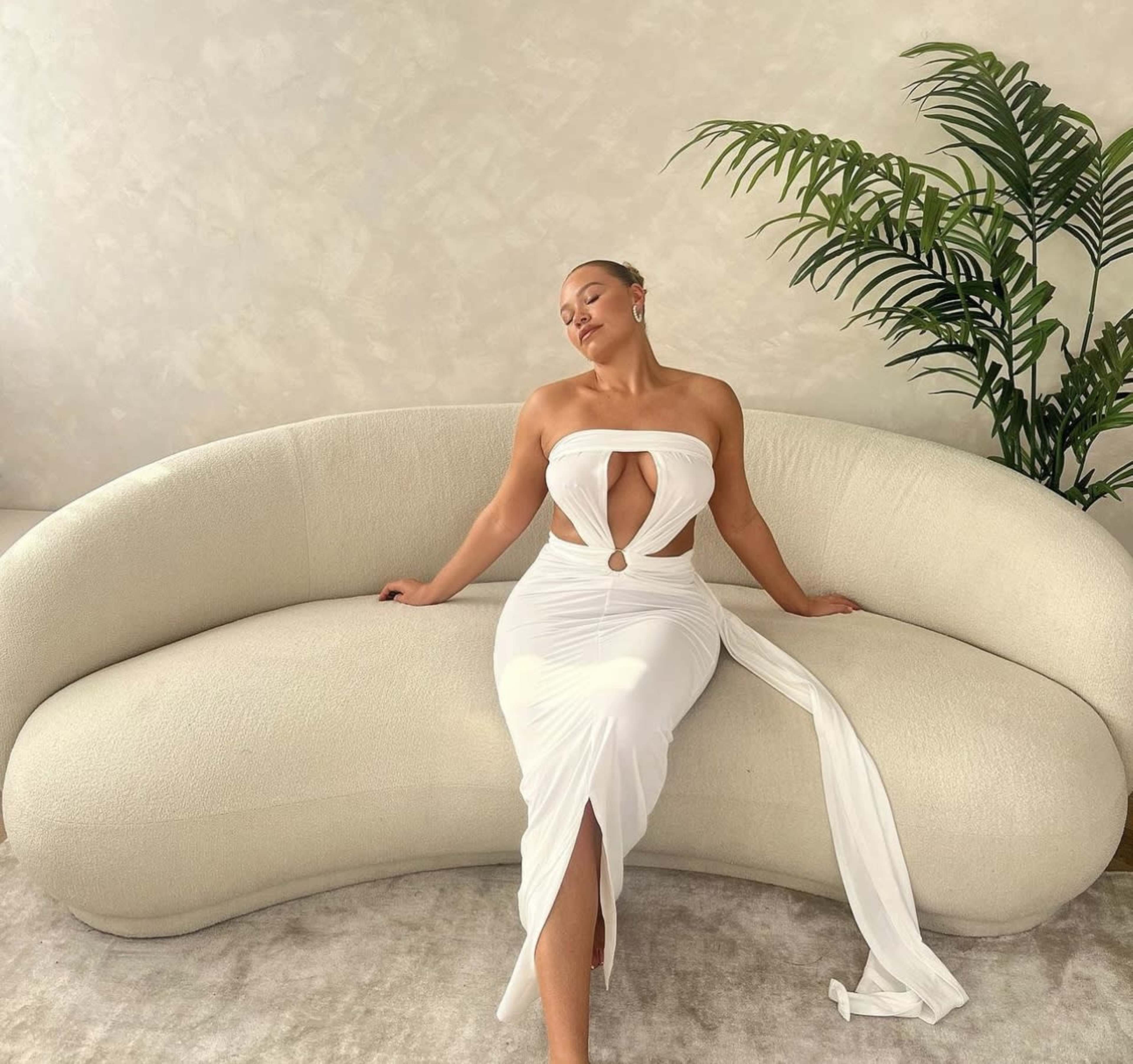 A woman in a white, strapless dress sits on a curved, neutral-toned sofa with a plant in the background.