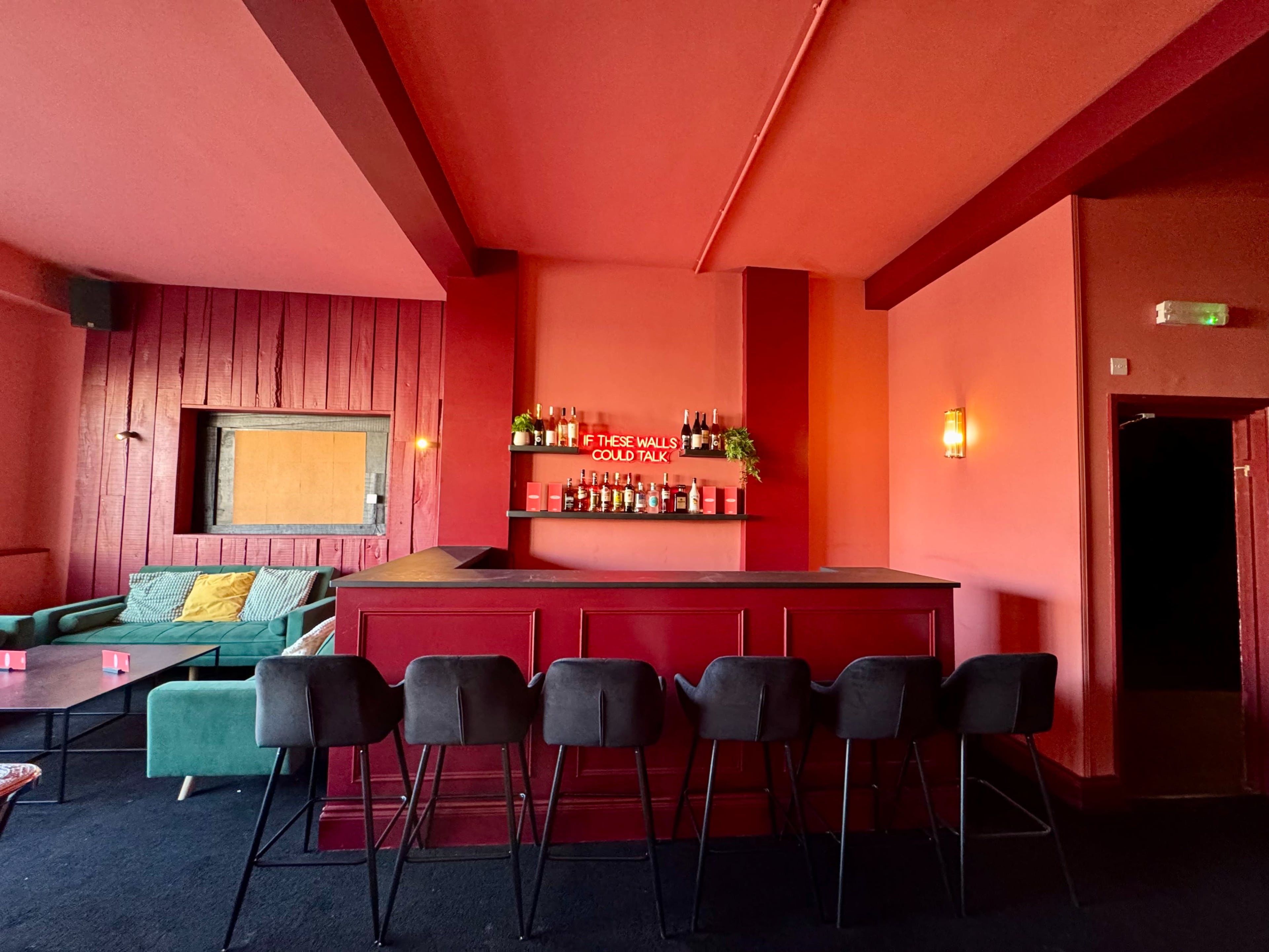 The image shows a bar area with a red interior, featuring a long counter, green couches, and a neon sign that reads "If these walls could talk."