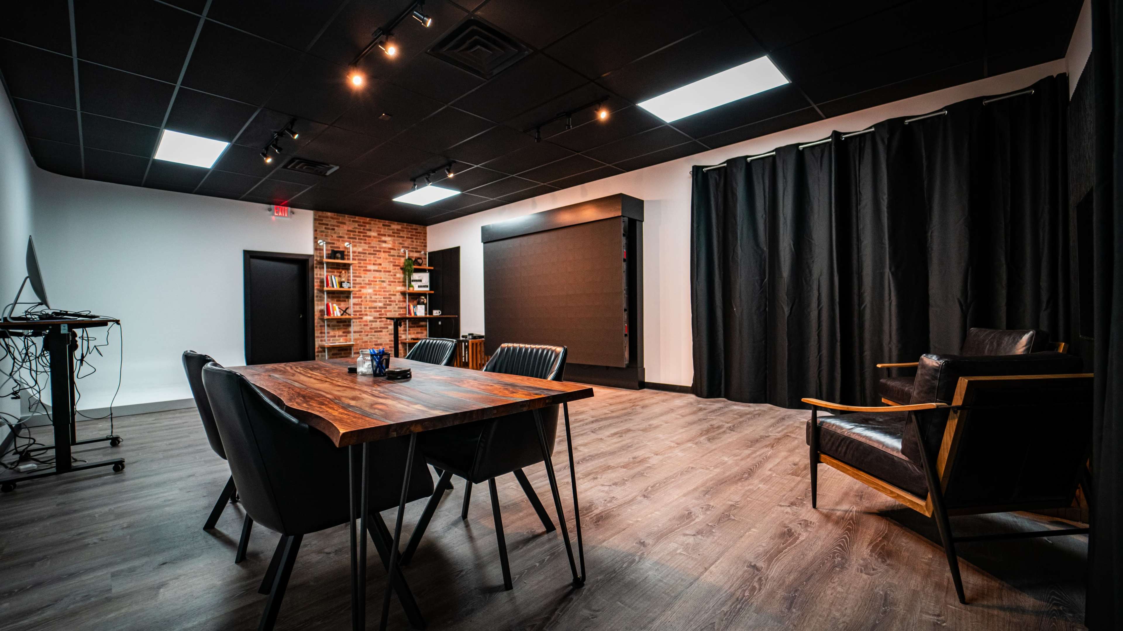A modern meeting room with a wooden table, black chairs, a bookshelf, and a dark wall with curtains.
