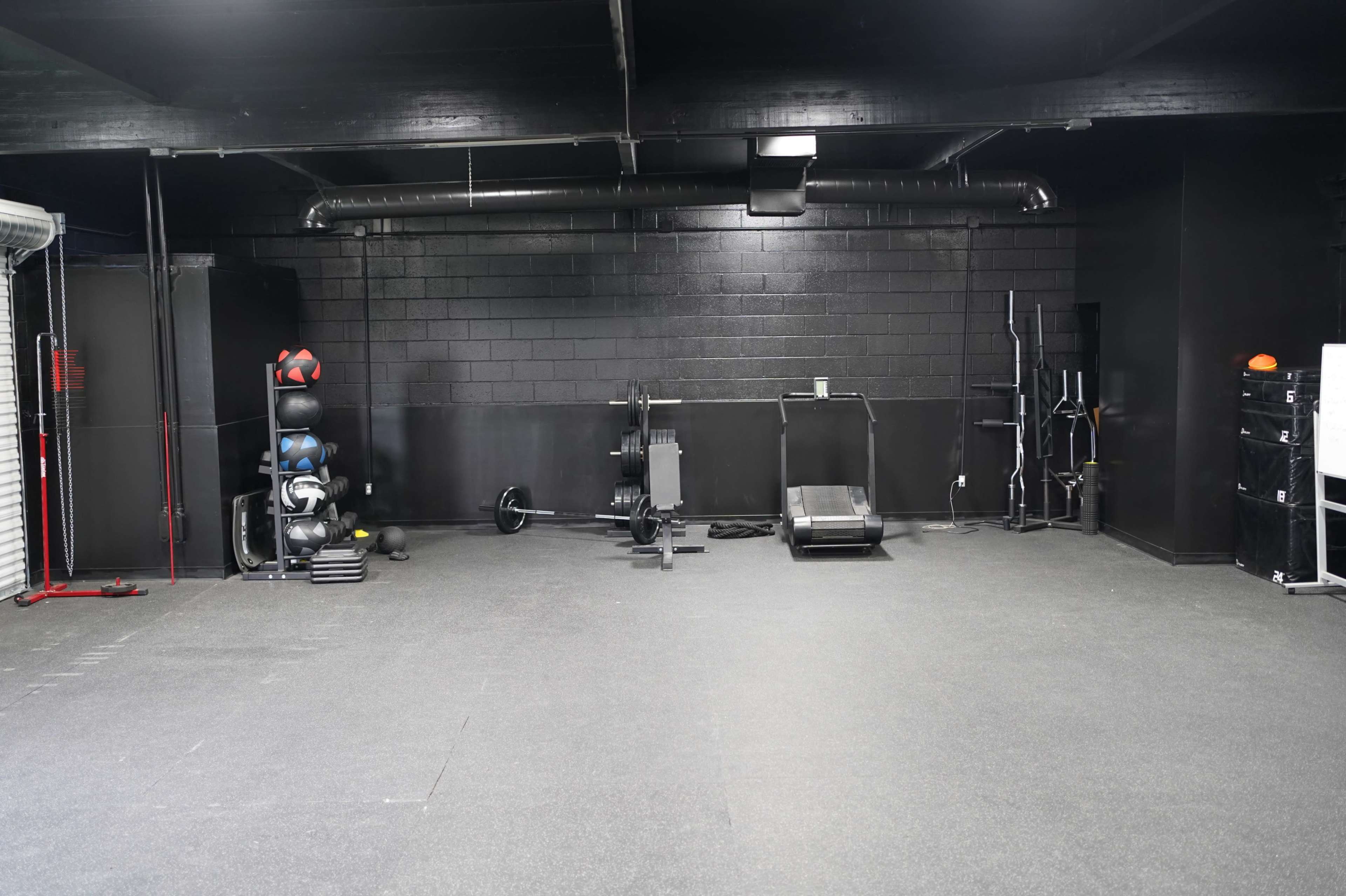 An empty black-walled gym with various exercise equipment, including weights, a treadmill, and a sled on a rubber flooring surface.