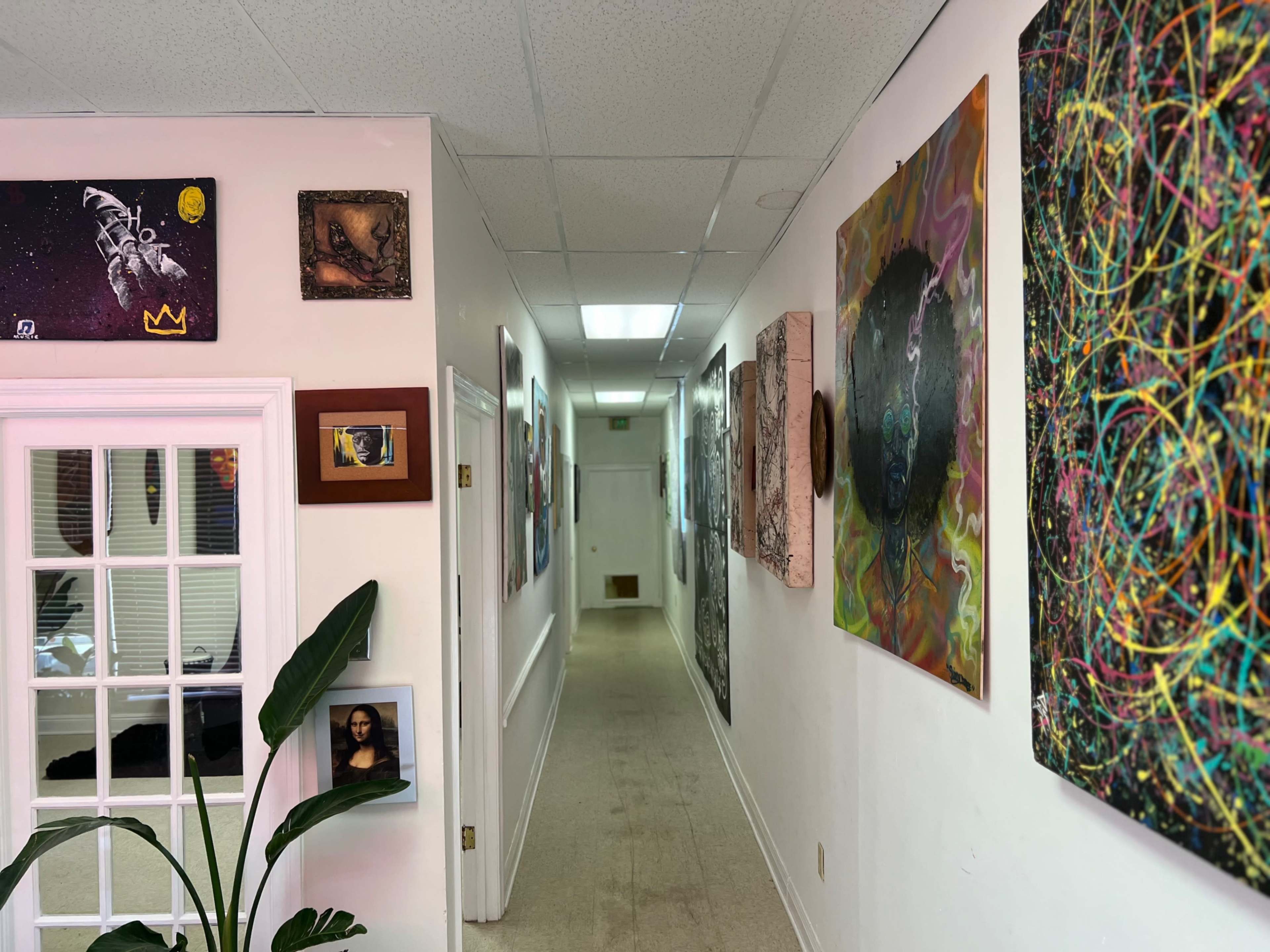 A corridor with white walls adorned with various pieces of art, leading to a brightly lit doorway at the end.