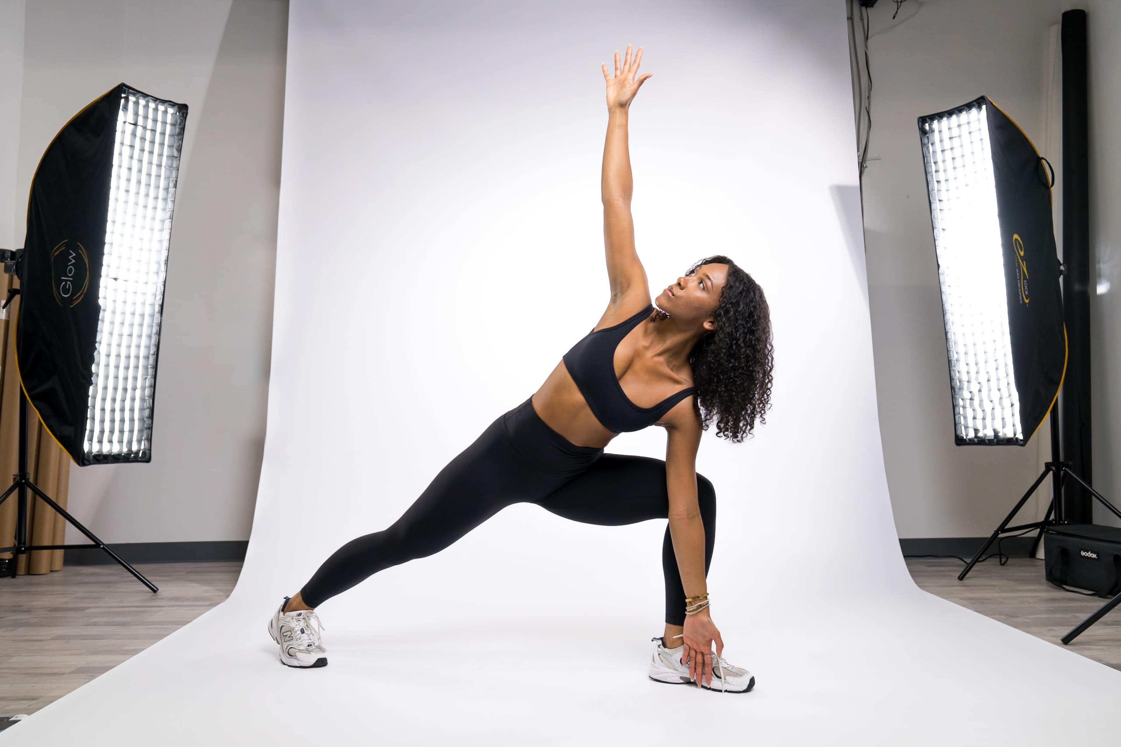 A woman in a black crop top and leggings performs a stretching pose on a white backdrop, illuminated by two lights.