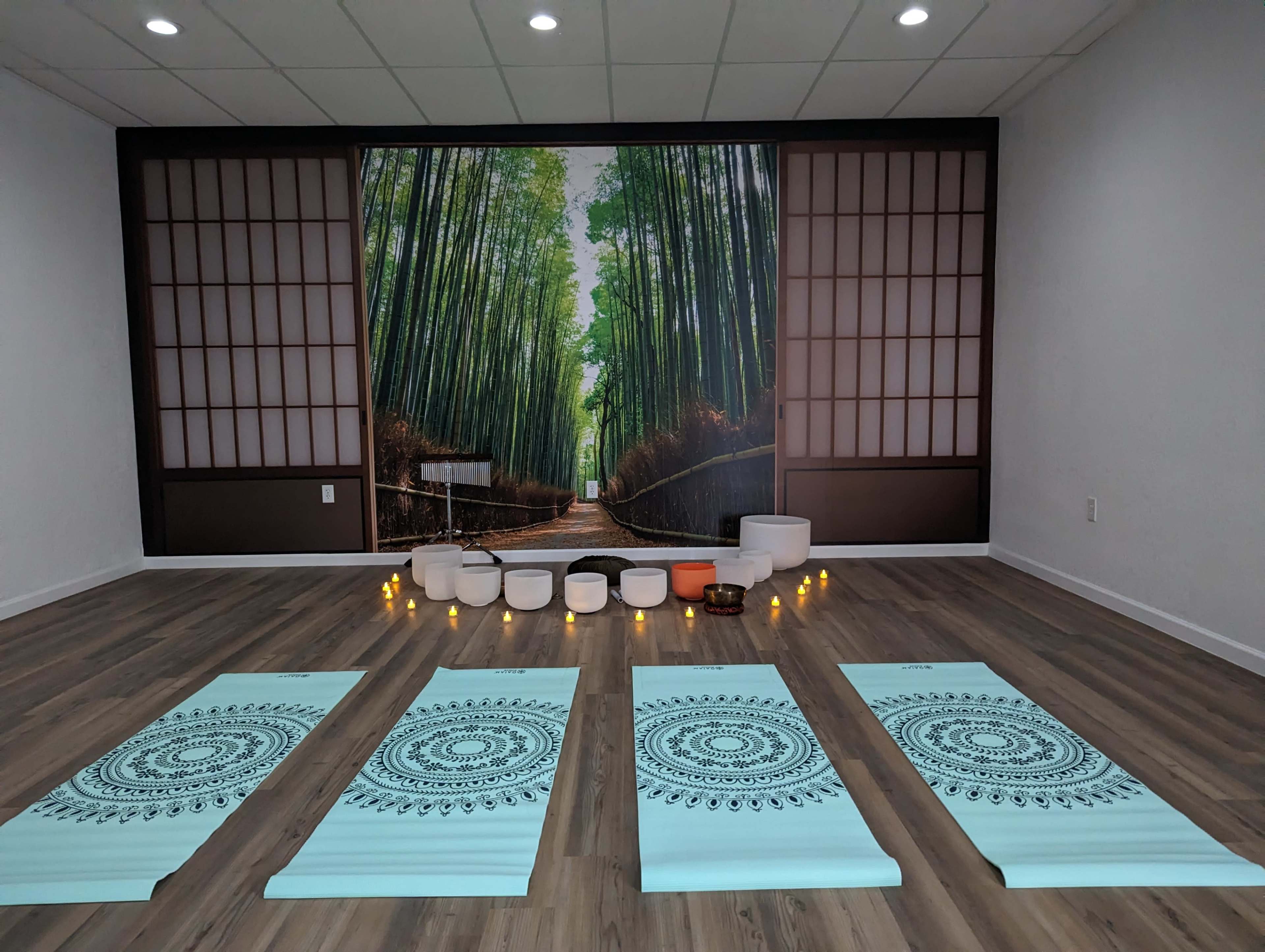 A yoga studio featuring four yoga mats arranged in a row, with a backdrop of a forest mural and bowls set up at the front, surrounded by candles.