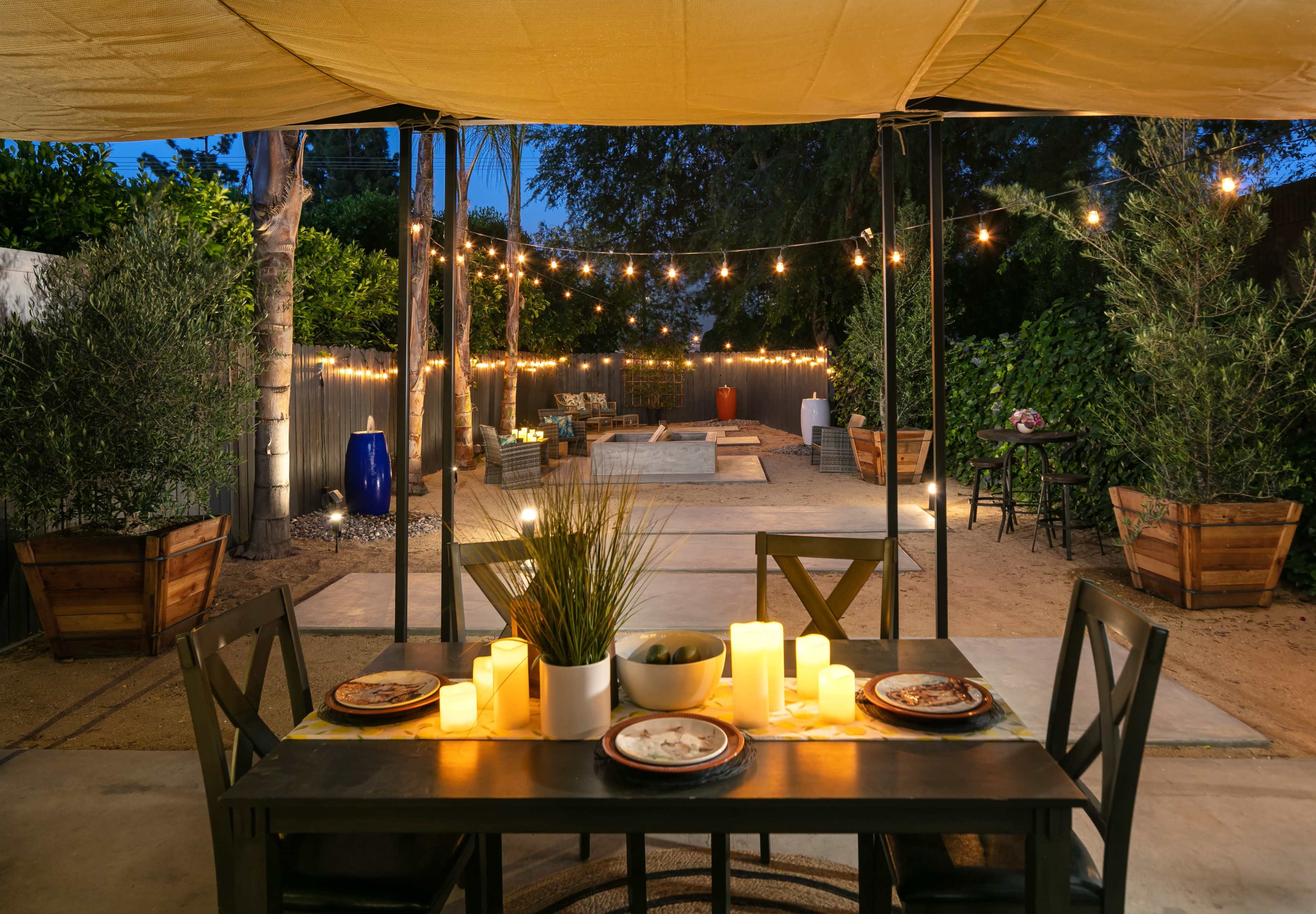 A table set with plates and lit candles is positioned under a canopy, overlooking a backyard adorned with string lights and potted plants.