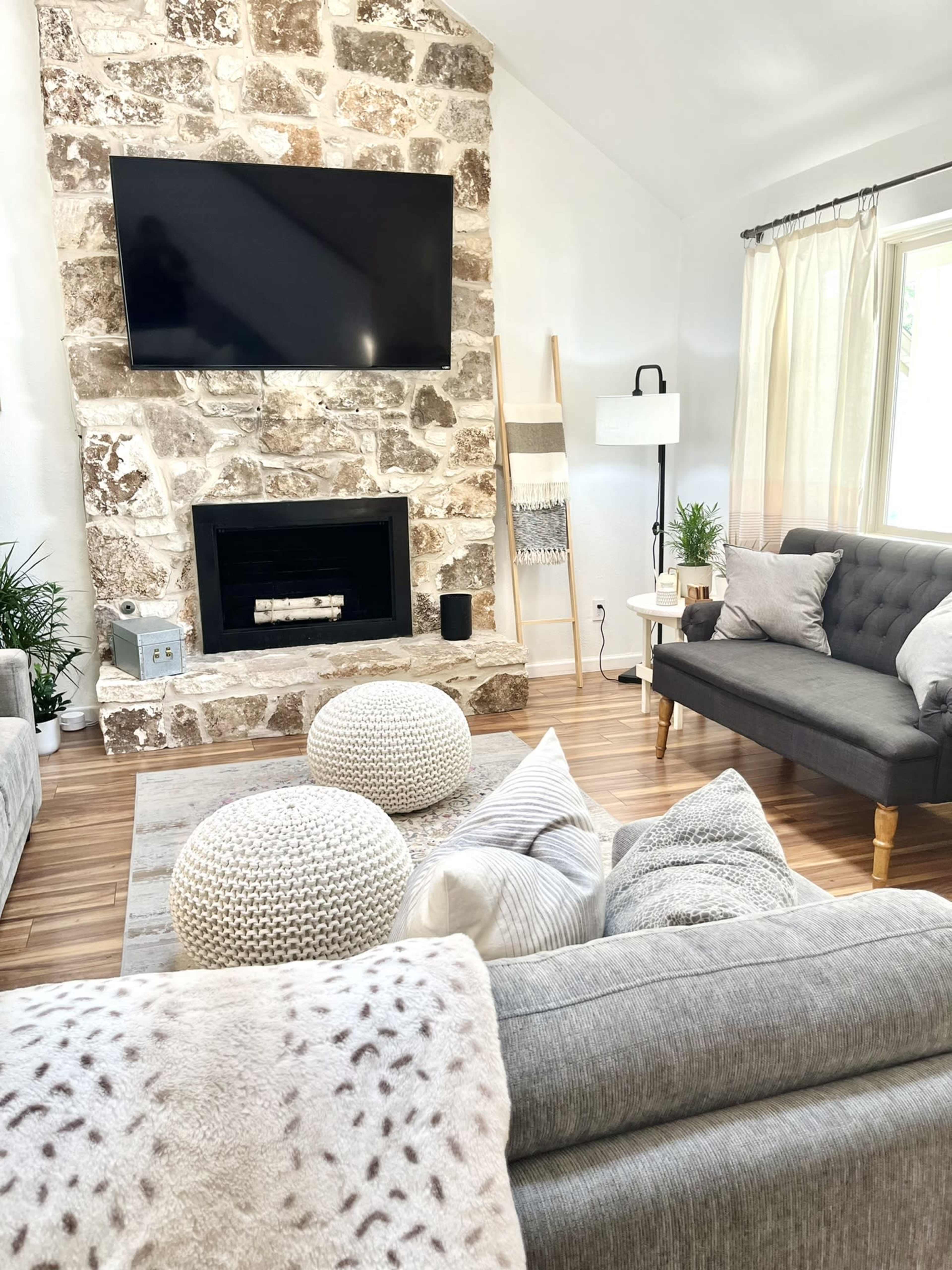The living room features a stone fireplace with a mounted TV, a gray sofa, a light-colored armchair, and decorative poufs on a wooden floor.