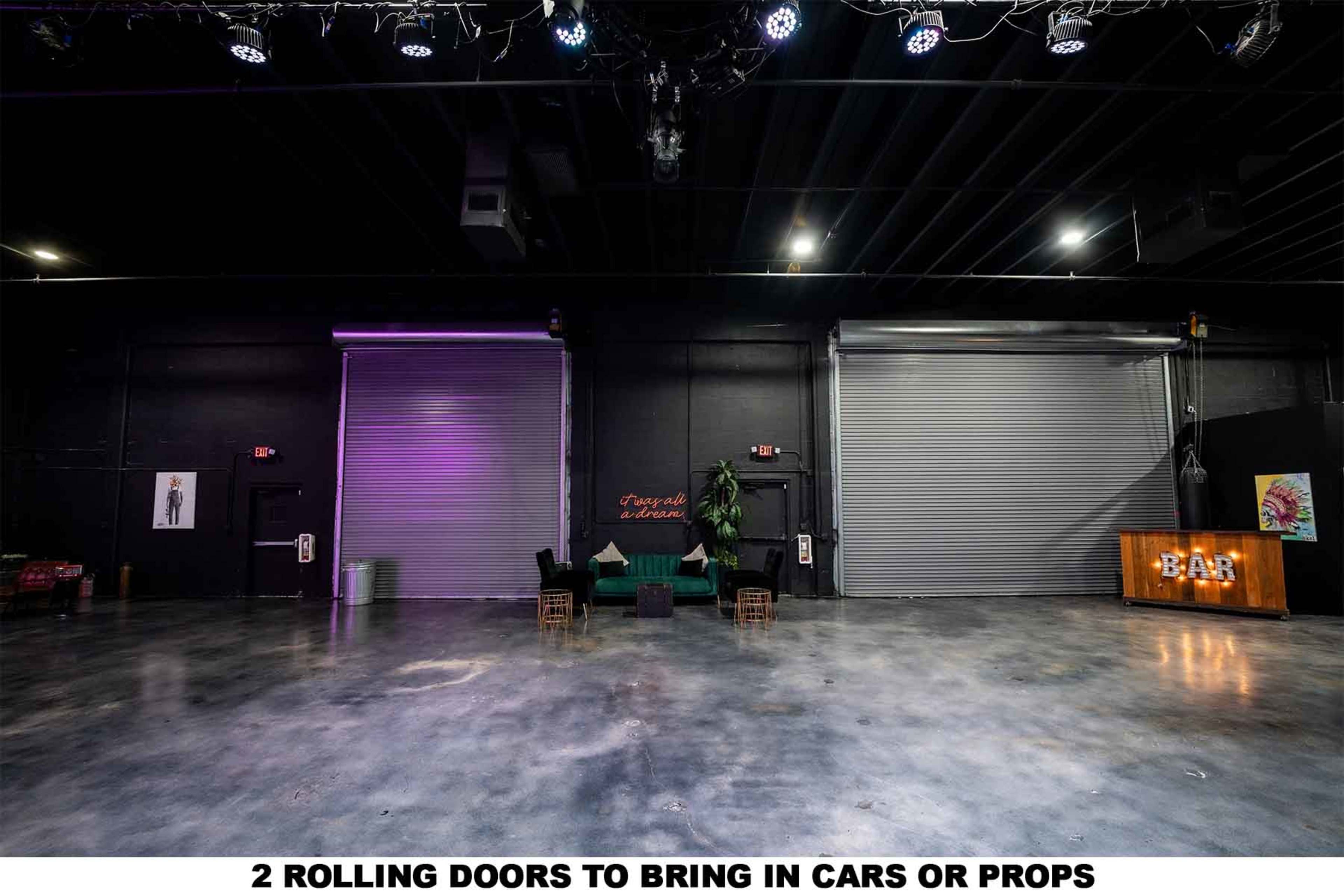 The image shows a spacious indoor venue with two large rolling doors on one side, a bar area, and minimal furnishings.