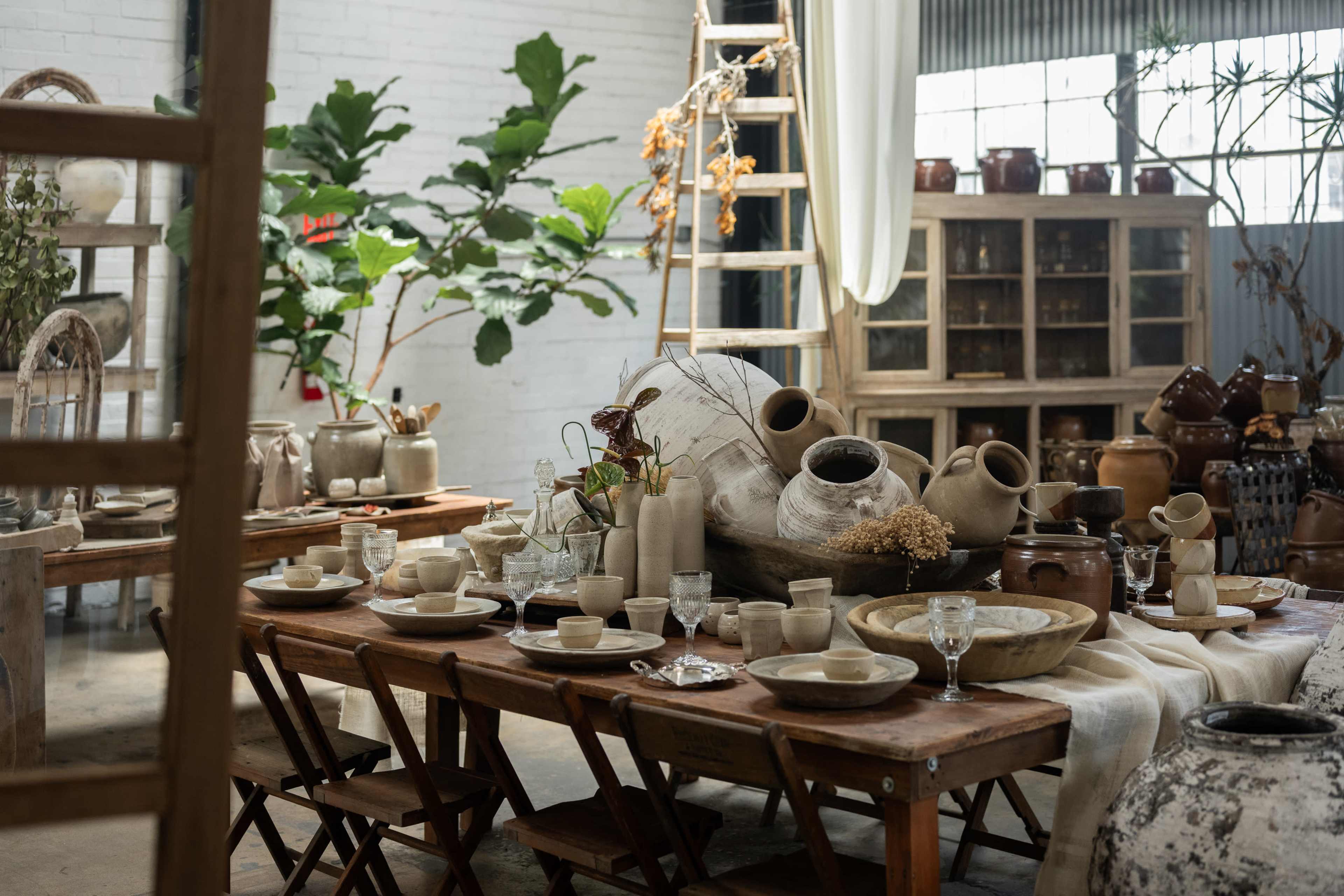 The image shows a rustic interior filled with pottery and tableware arranged on wooden tables, surrounded by greenery and shelves of clay jars.
