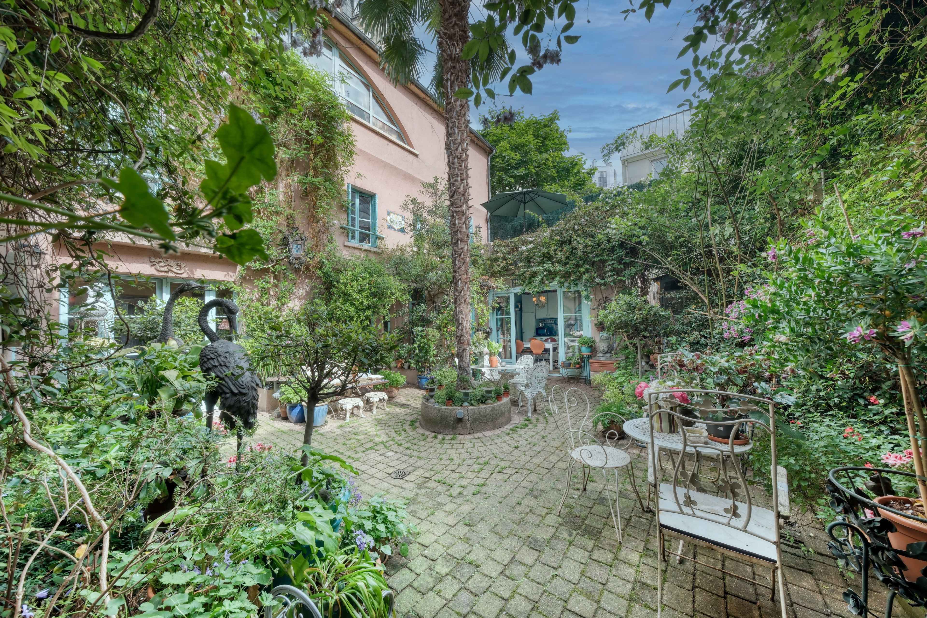 The image depicts a lush garden courtyard surrounded by a pink building, featuring various plants, outdoor seating, and a sculpture.