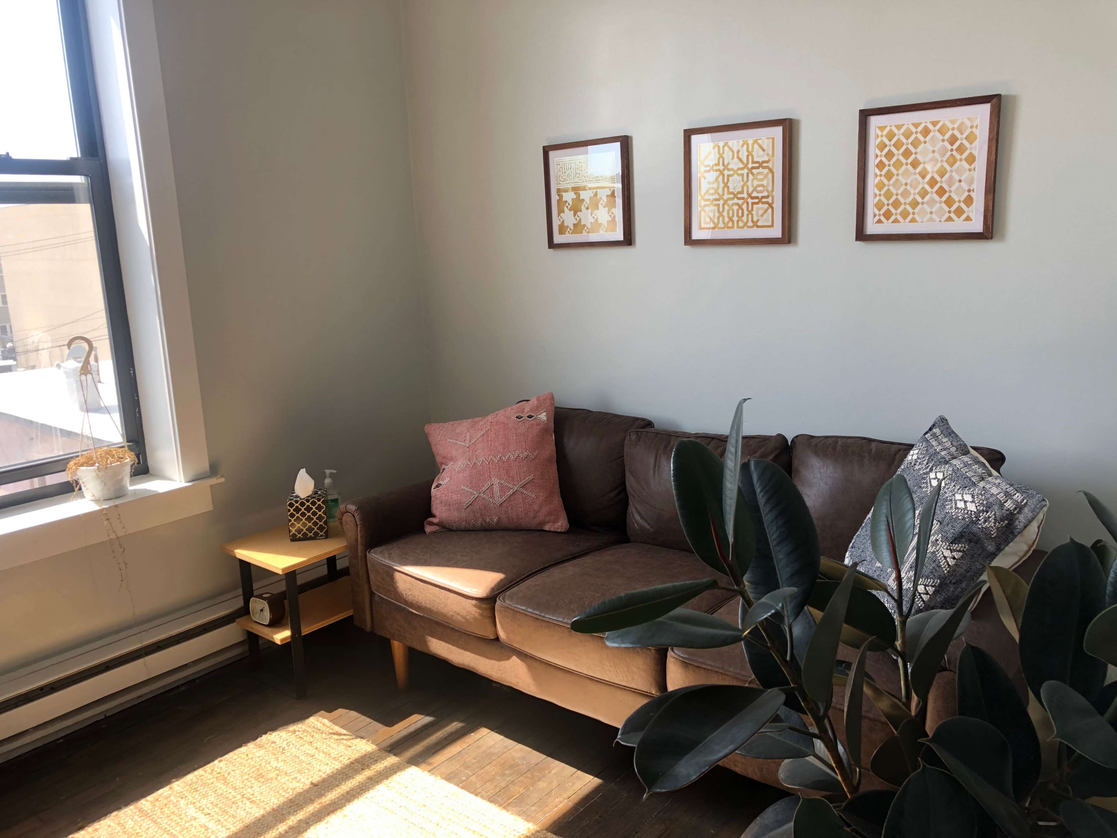 A brown couch with decorative pillows sits in a bright corner of a room, accompanied by a small side table and three framed artworks on the wall.