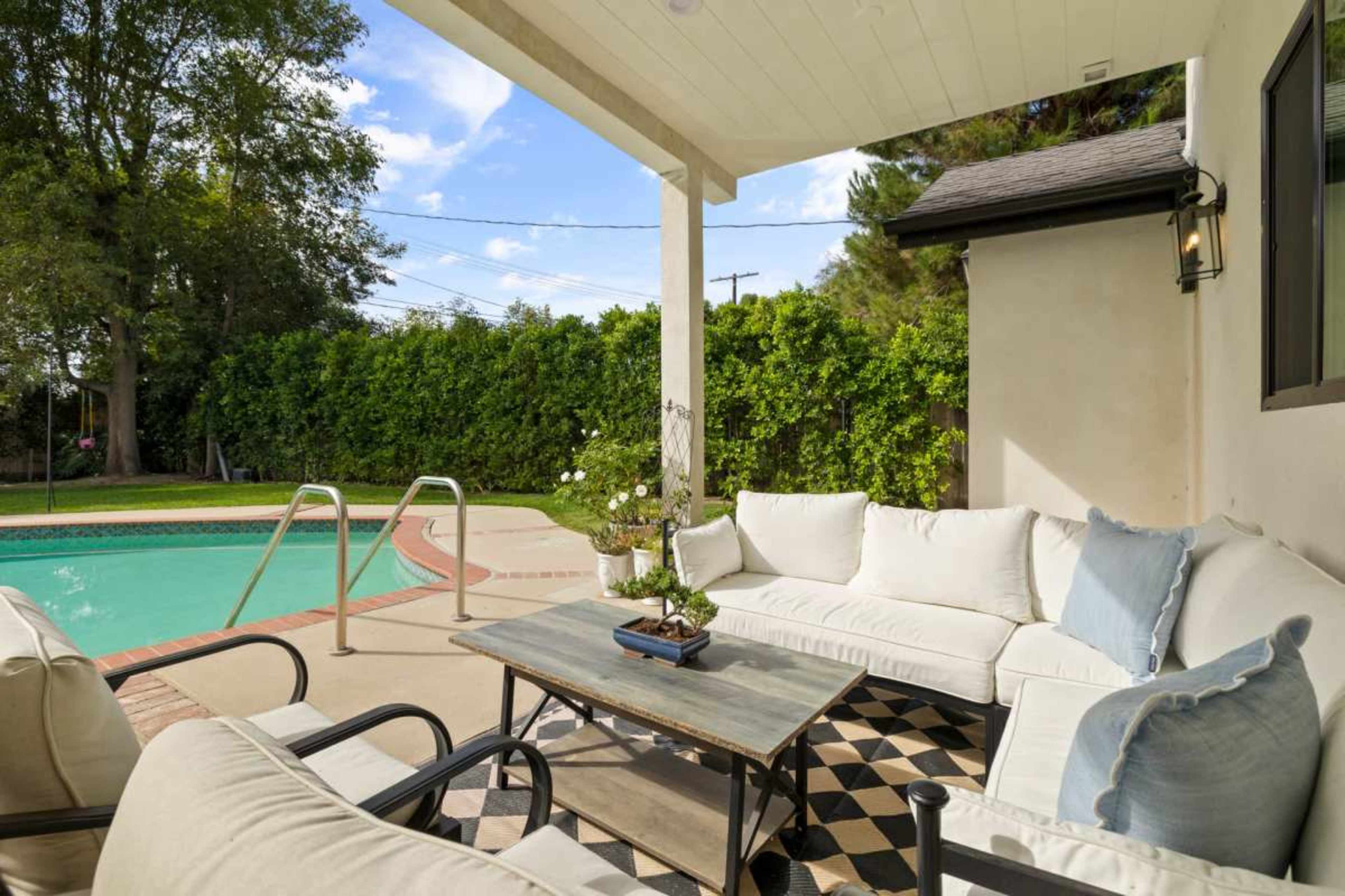 The scene features an outdoor seating area with a sectional sofa, a coffee table, and a swimming pool in the background surrounded by greenery.