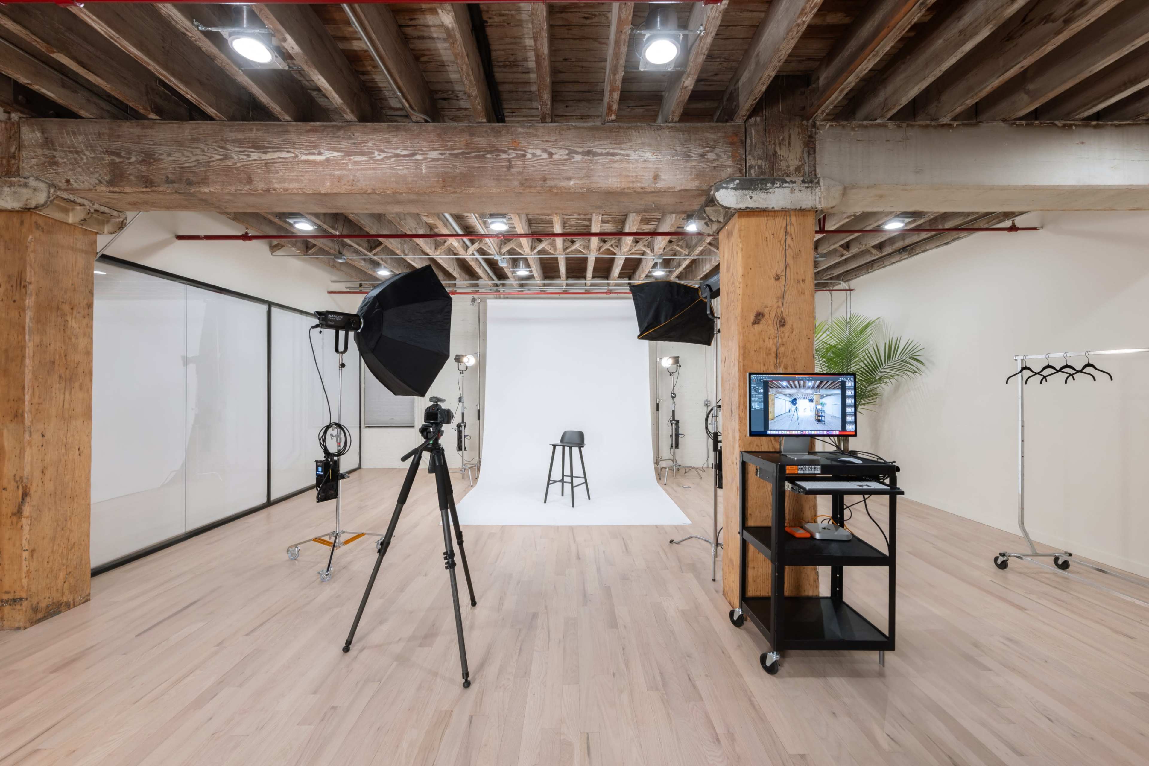 A photography studio features a white backdrop, a stool, lighting equipment, a camera on a tripod, and a monitor on a mobile cart.