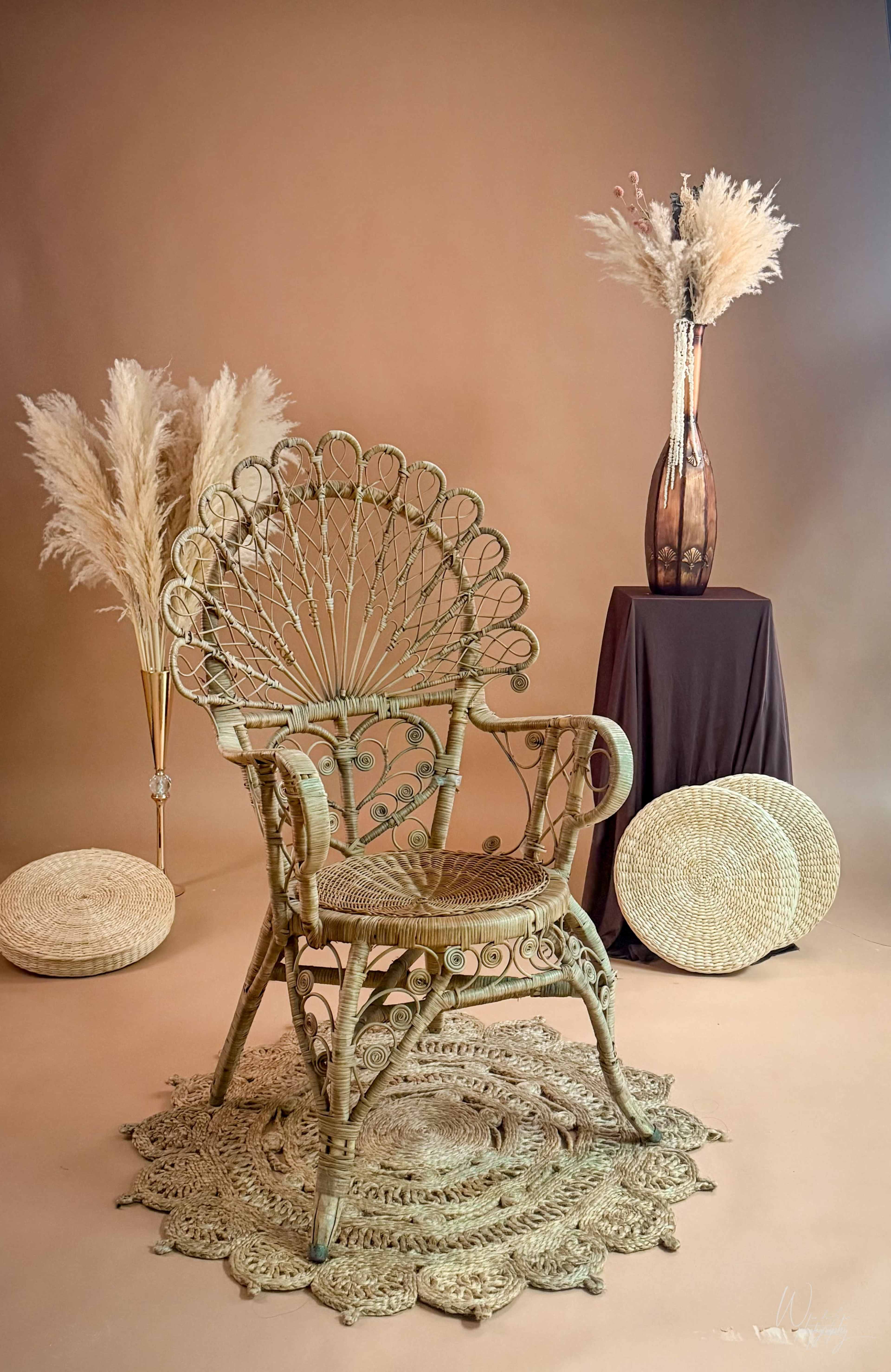 The image features a decorative chair made of woven materials, surrounded by dried pampas grass in a vase, a dark tablecloth-covered pedestal, and circular woven mats on the floor.