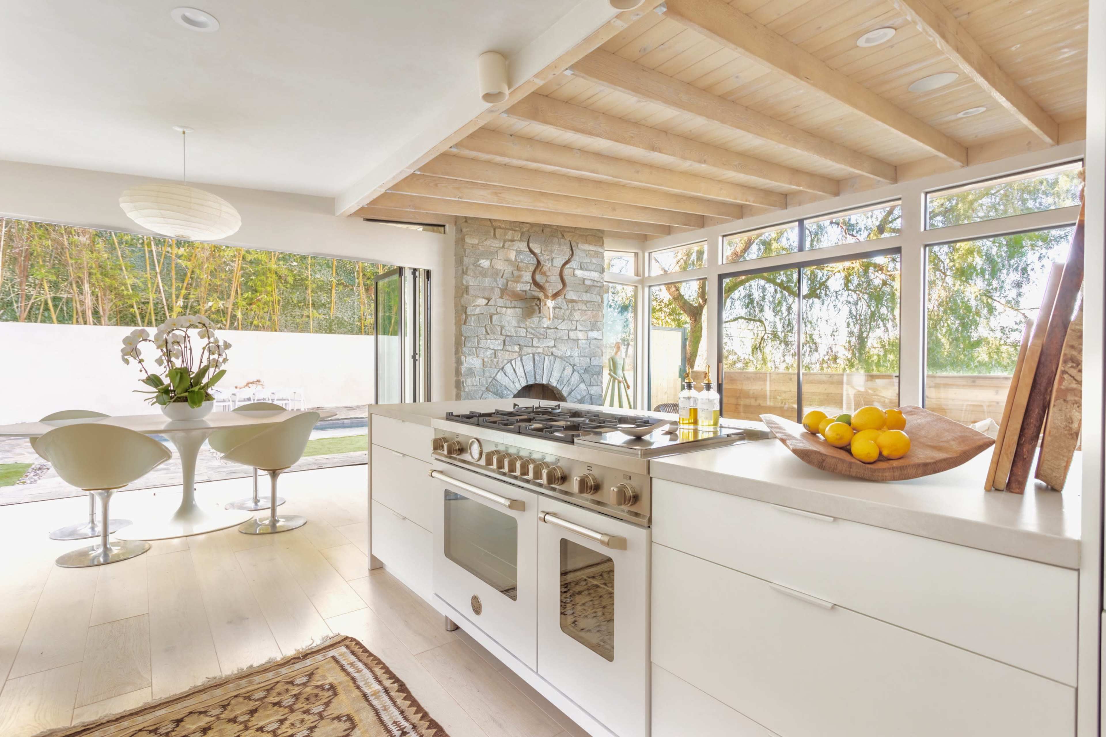 A bright kitchen features a gas stove, white cabinets, and a dining area with a round table surrounded by modern chairs, overlooking a garden through large windows.